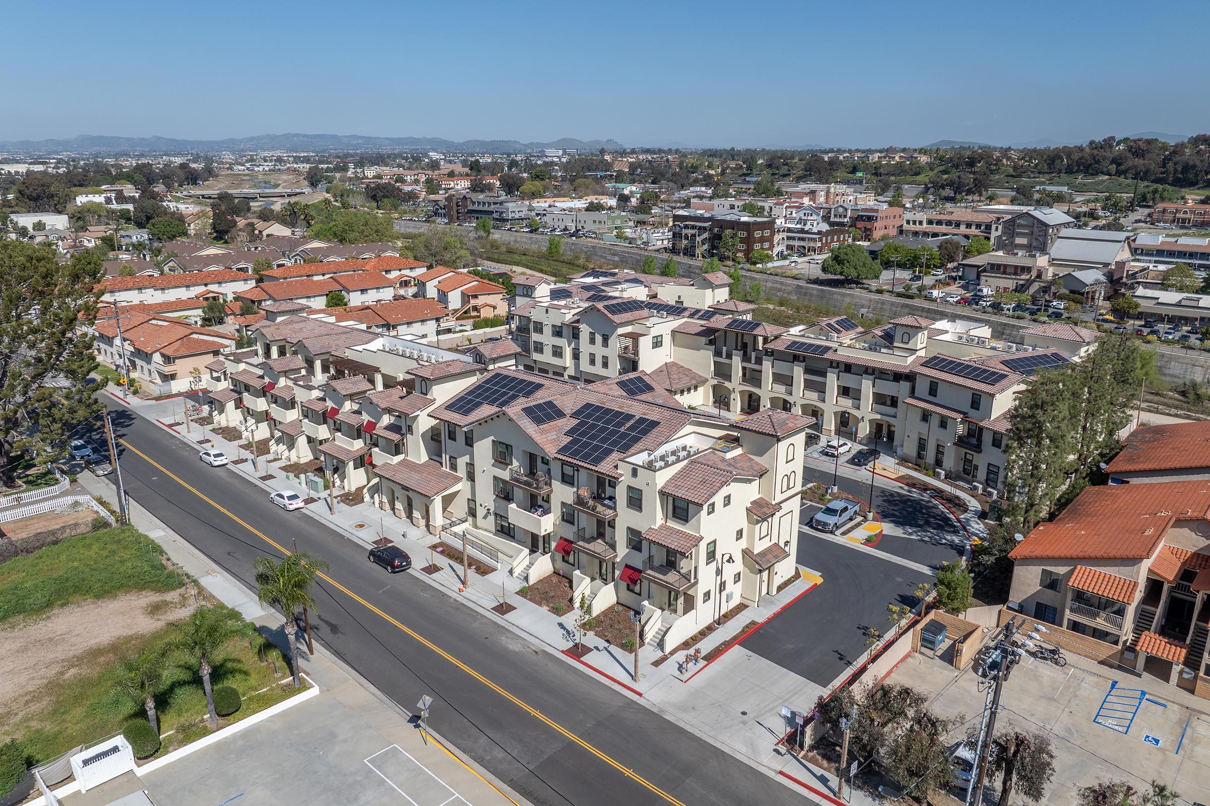 Aerial view of a modern residential neighborhood featuring multi-story buildings with solar panels on the roofs. The streets are lined with palm trees and parked cars, with additional houses nearby. The background includes a lush landscape and distant hills under a clear blue sky.