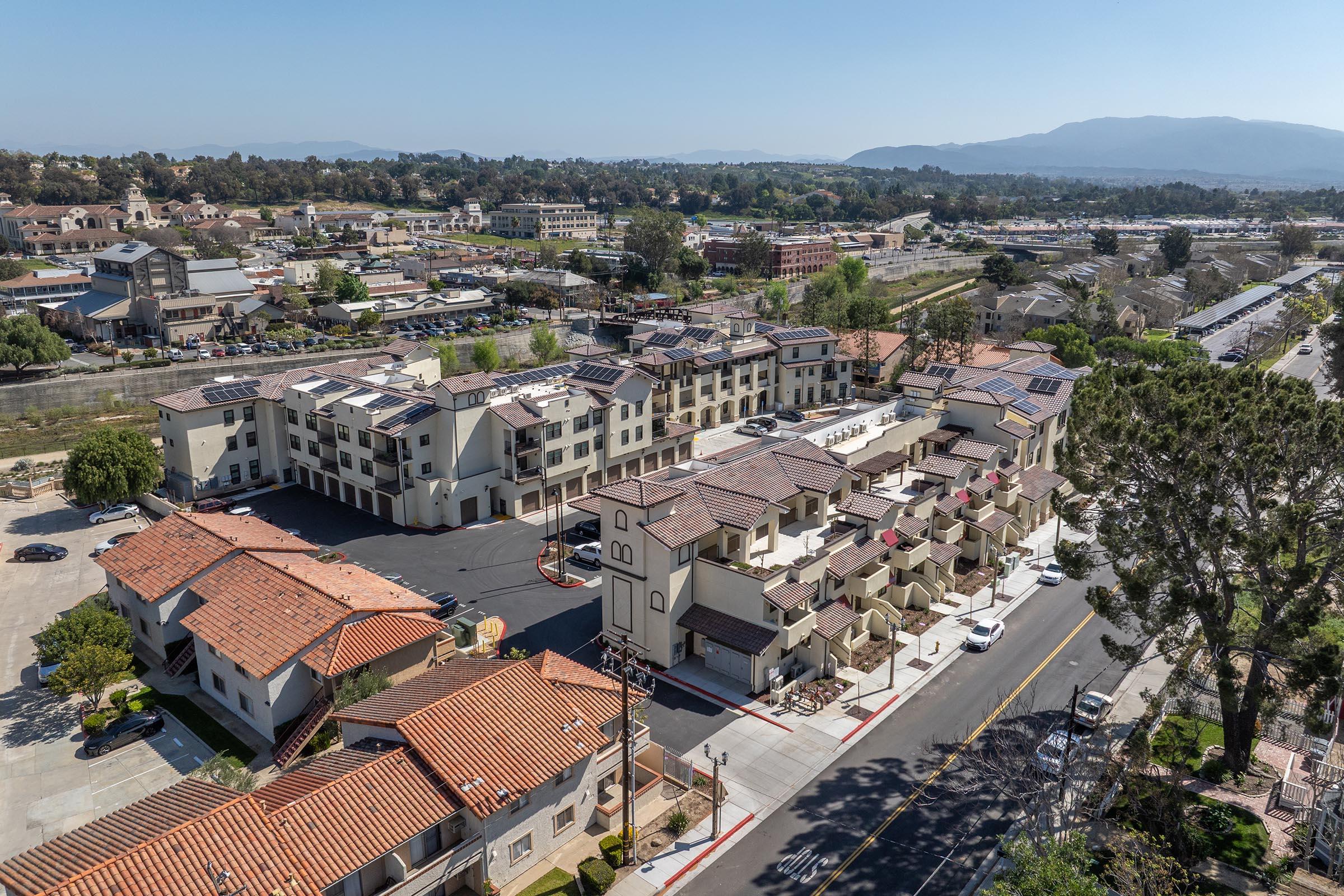 Aerial view of a residential area featuring a multi-story building with a tiled roof, surrounded by smaller homes and green spaces. In the background, more buildings and mountains are visible along with roads and trees, showcasing a suburban landscape with a mix of architecture.