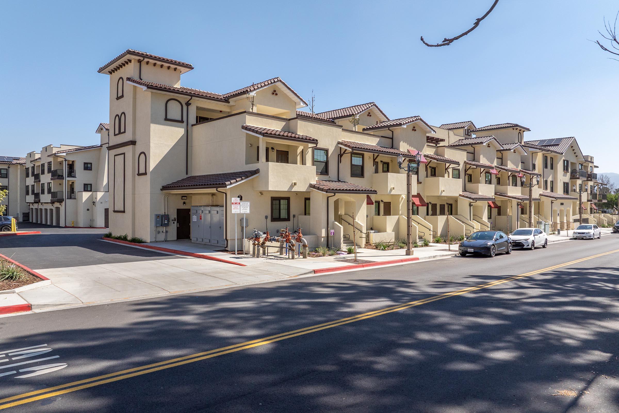 A row of beige-colored multi-story residential buildings with a Spanish-style architectural design, featuring balconies and tiled roofs. The street in front has a sidewalk, parked bicycles, and a couple of cars. Trees cast shadows on the pavement, creating a sunny urban environment.