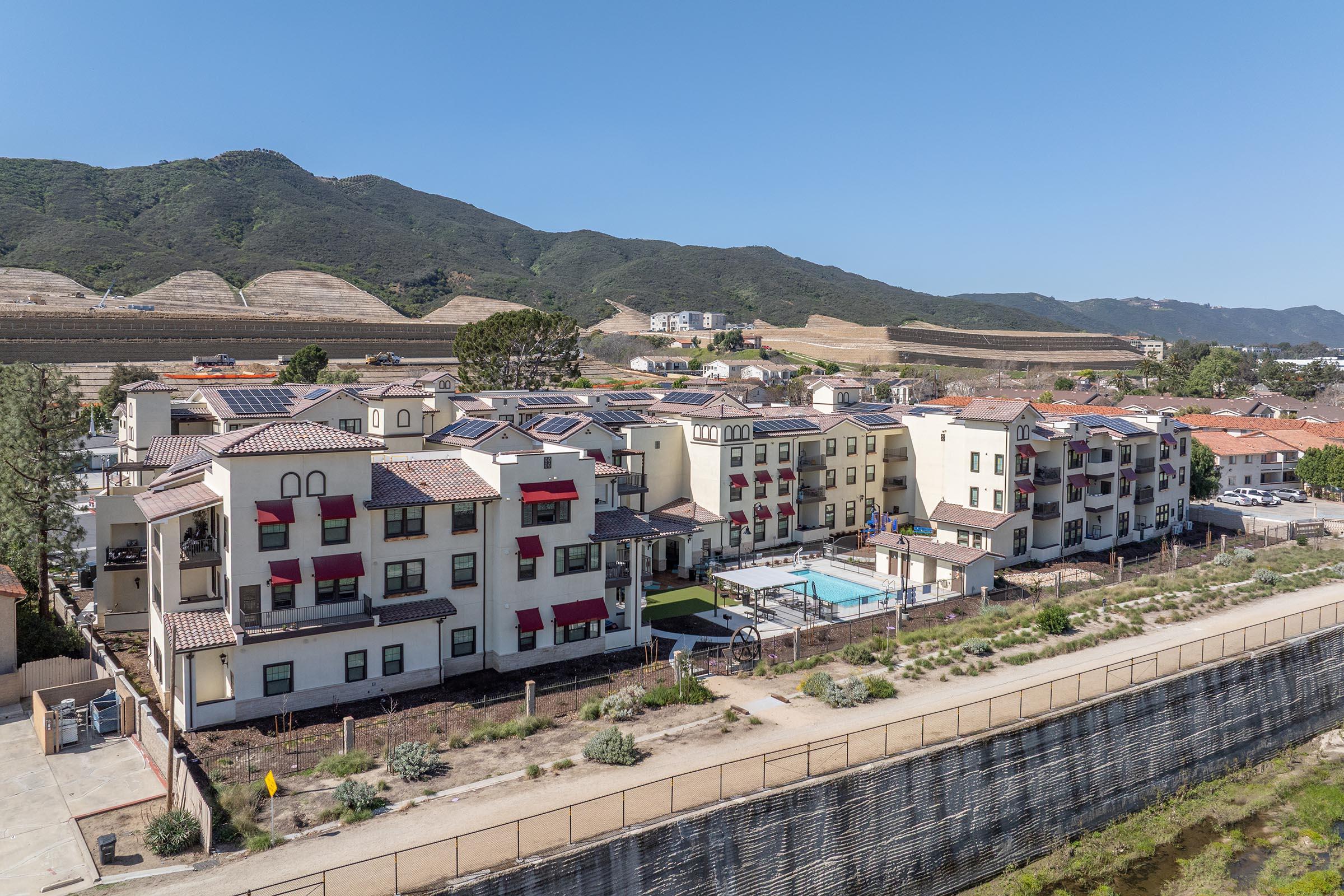 Aerial view of a modern apartment complex with a pool, surrounded by landscaped greenery. In the background, hills are visible, along with other residential buildings. The clear blue sky enhances the sunny atmosphere of the scene.