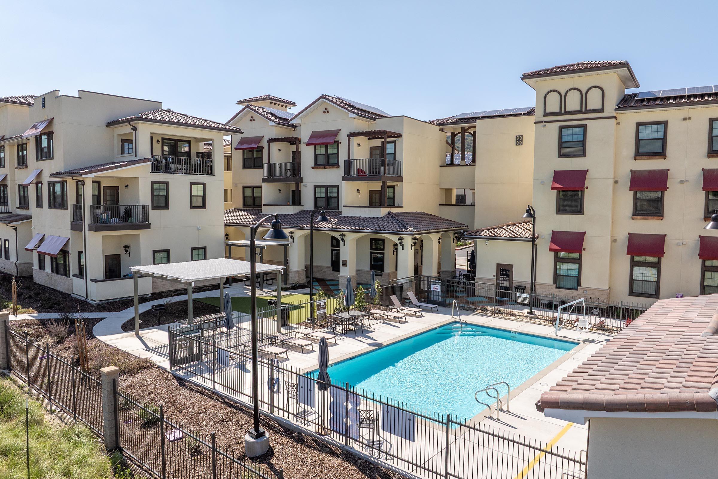 An aerial view of an apartment complex featuring a pool area. The pool is surrounded by lounge chairs and shaded by awnings, with several buildings in the background. The architecture includes multiple levels, balconies, and decorative elements, all set in a sunny, landscaped environment.