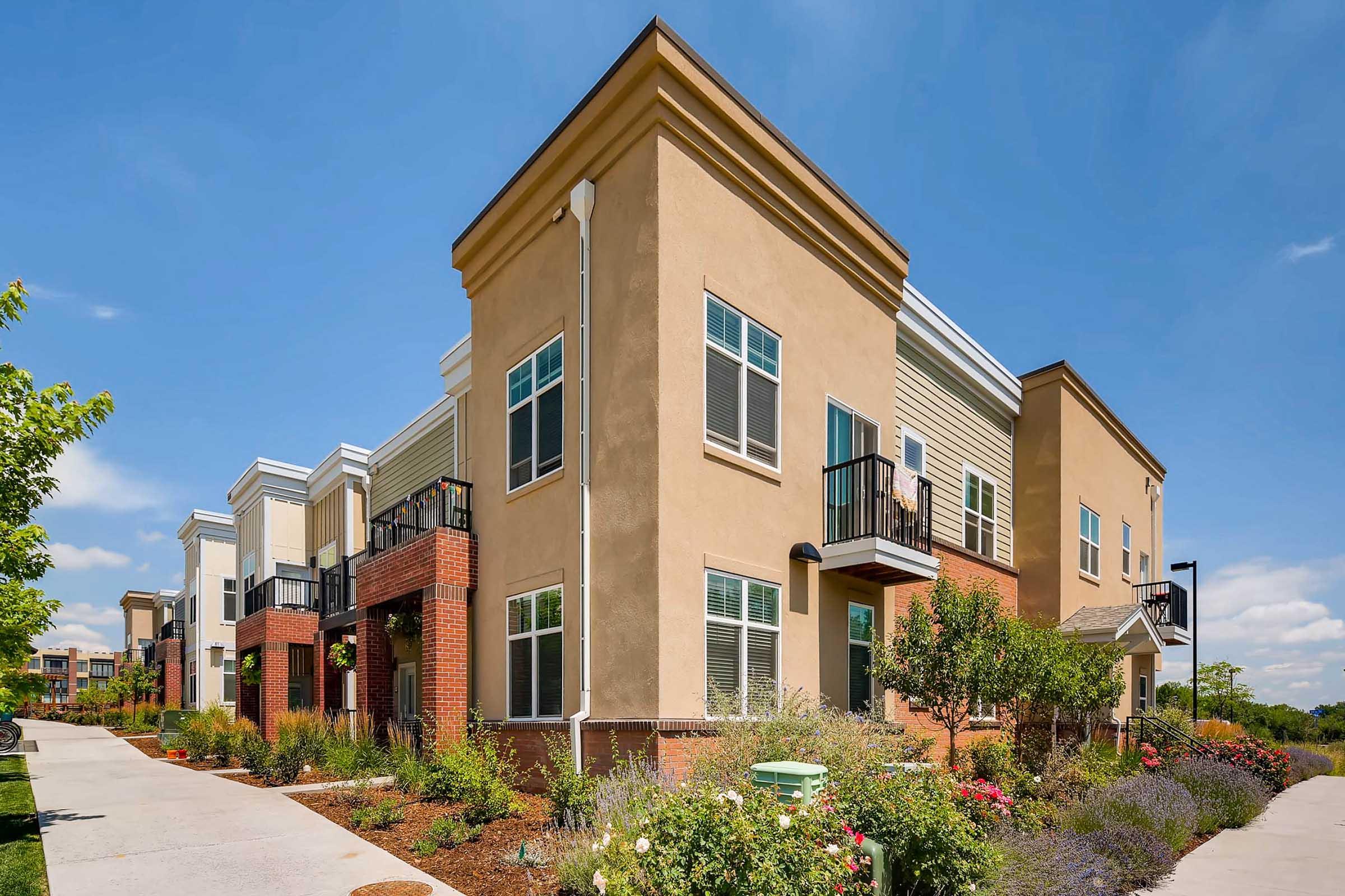 Modern residential building with a blend of light-colored stucco and brick facades, featuring balconies and surrounded by landscaped greenery. A concrete walkway leads through the garden area, under a clear blue sky.