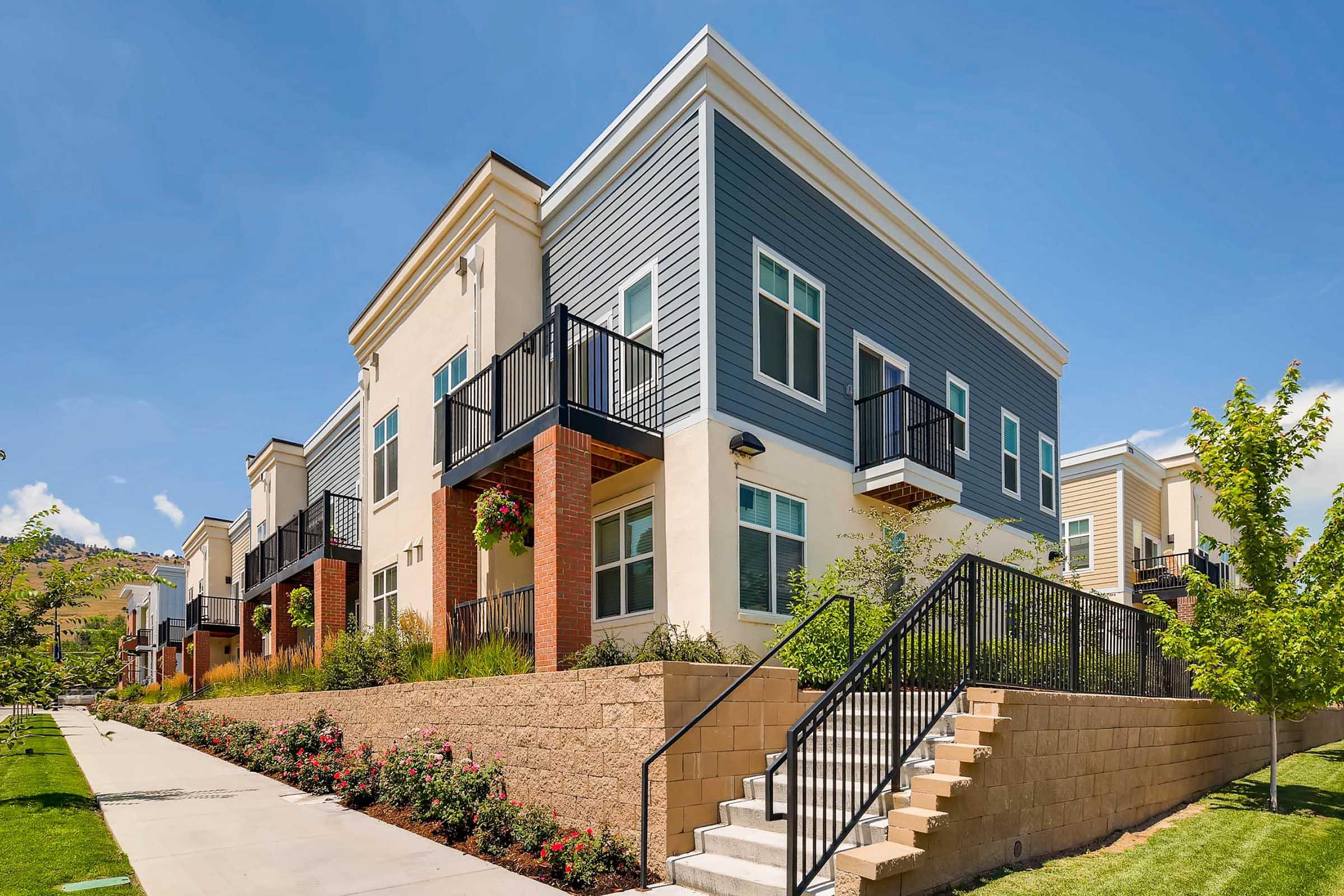Modern multi-unit residential building with a mix of light and dark siding. It features multiple balconies, landscaped gardens, and a pathway leading to a staircase. The sky is clear and blue, contributing to a bright, inviting atmosphere around the property.