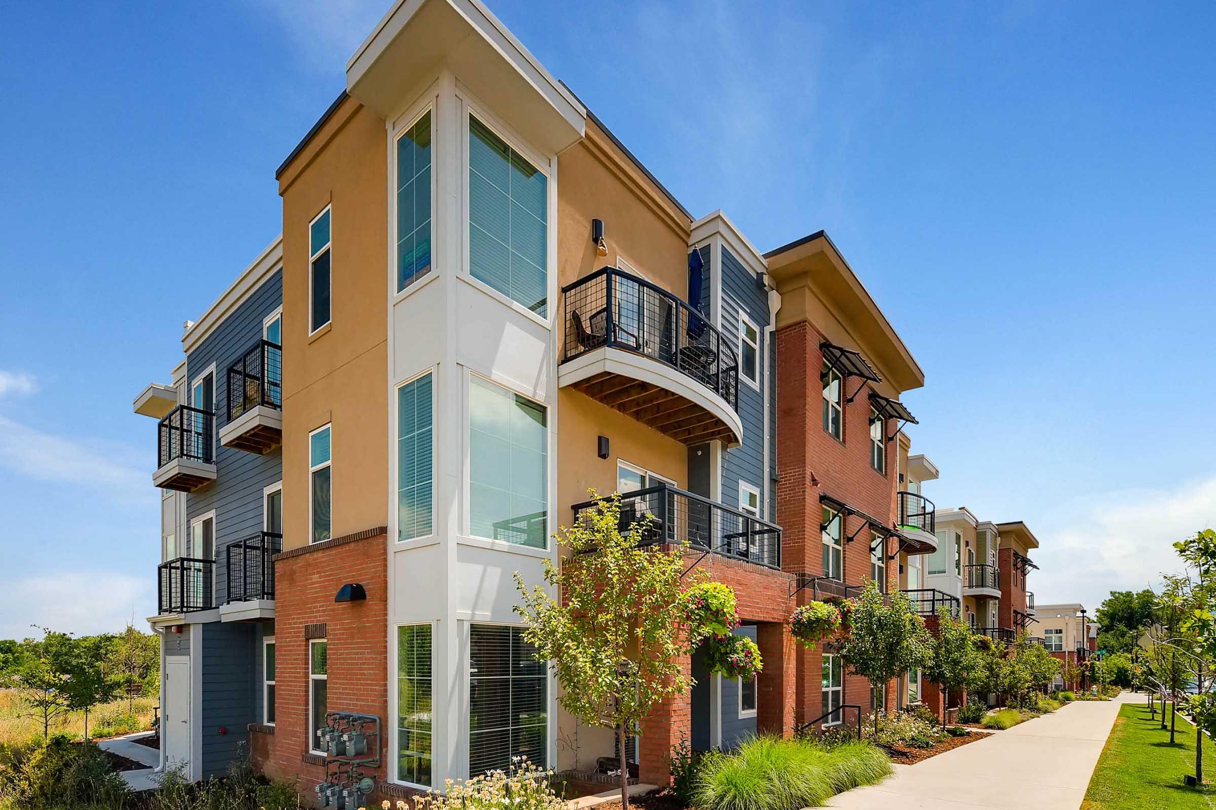 Modern multi-unit apartment building with a mix of brick and stucco exteriors. Features large windows, balconies, and landscaped pathways lined with trees and shrubs. The setting is bright and sunny, with a clear blue sky in the background.