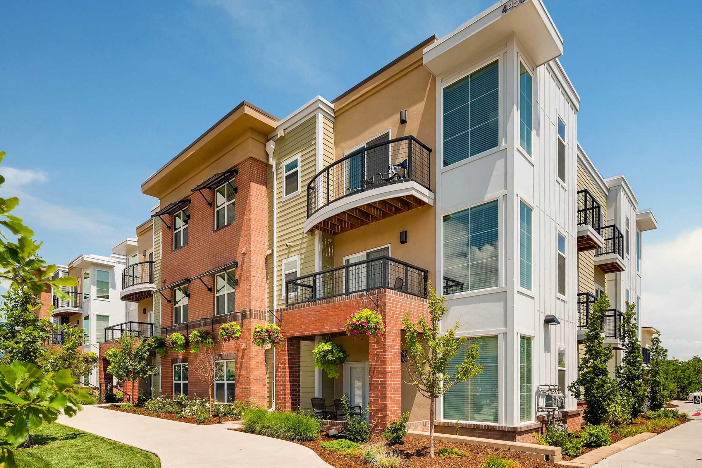Modern multi-story apartment building with a mix of brick and light-colored siding. Features include balconies, large windows, and landscaped pathways adorned with greenery and flower pots, set against a clear blue sky.