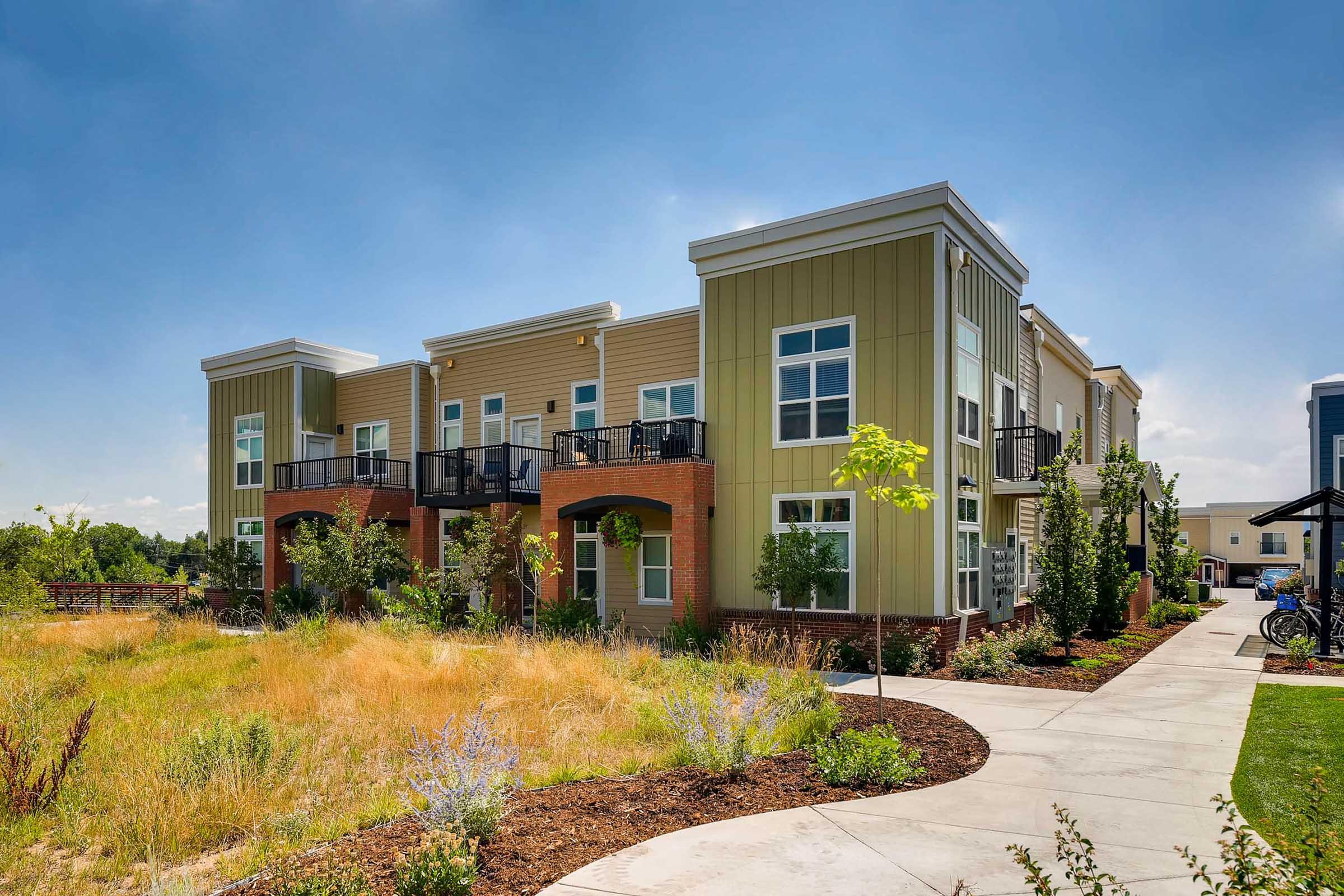 Two-story modern apartment building with balconies, surrounded by landscaped grounds featuring grass, small trees, and flowering plants. A paved walkway leads to the entrance, and there are bicycles parked nearby, under a clear blue sky.