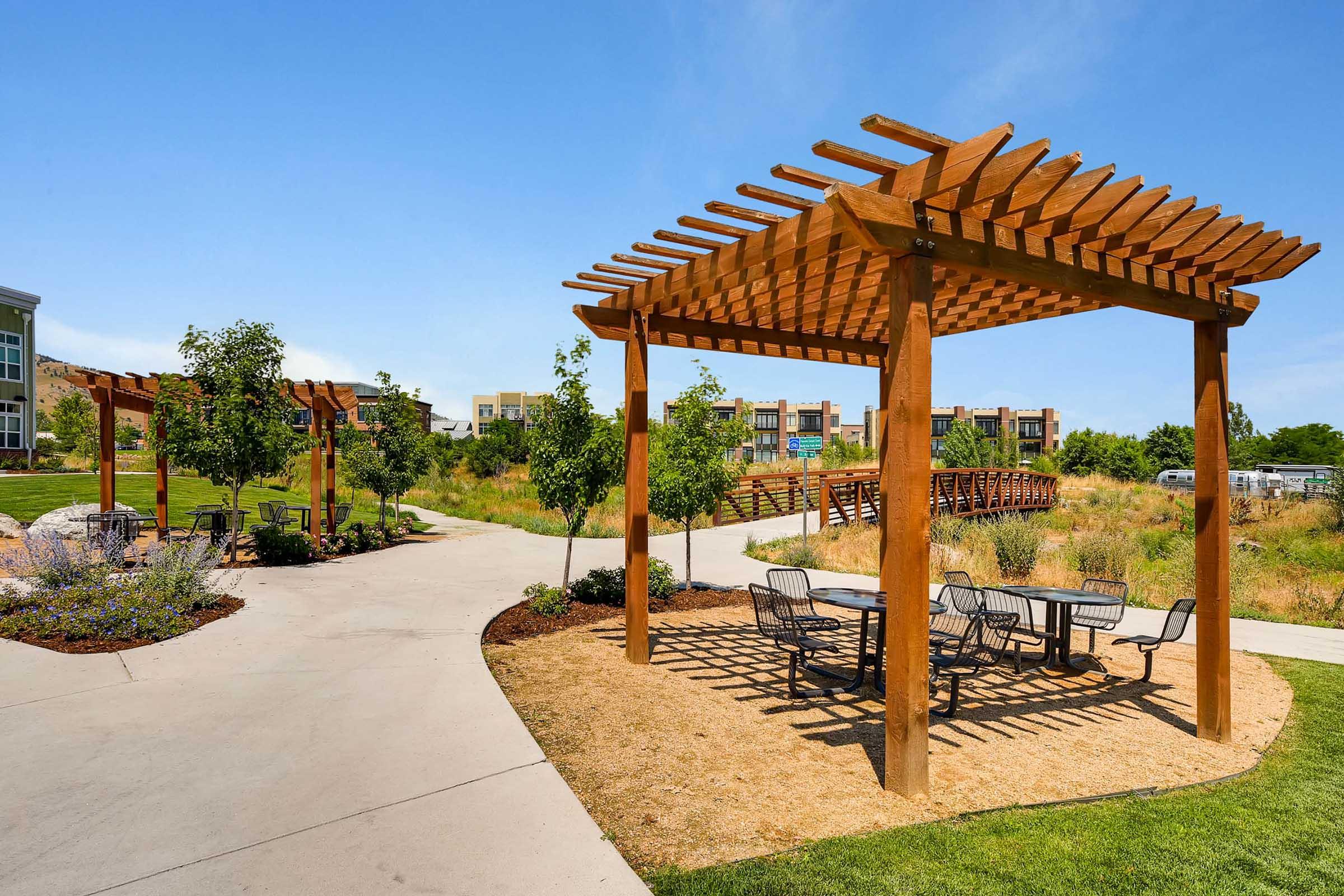 A sunny park scene featuring wooden pergolas with seating areas surrounded by lush greenery. A winding path leads into the background, where a wooden bridge is visible. The area is designed for relaxation and outdoor activities, showcasing a pleasant landscape with a clear blue sky.