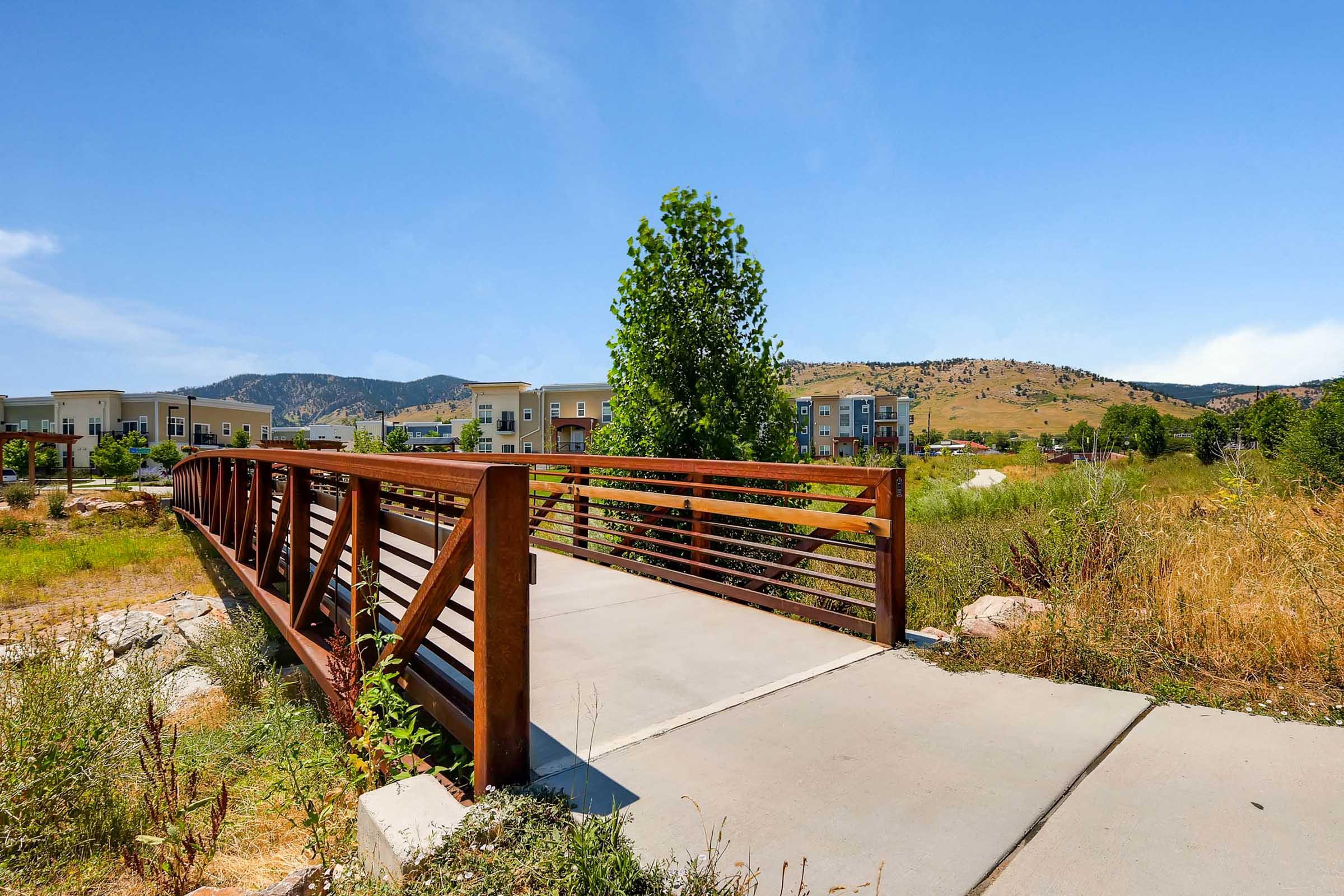 A metal pedestrian bridge crosses a pathway in a scenic outdoor area. Lush greenery and trees flank the bridge, with a backdrop of rolling hills and blue skies. Modern residential buildings are visible in the distance, adding a contemporary touch to the natural landscape.
