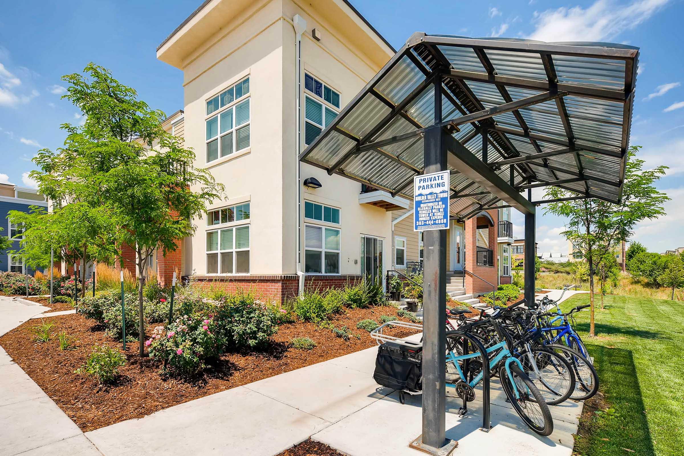 Bicycle parking area with a covered shelter, surrounded by landscaping featuring shrubs and small trees. In the background, a modern building with large windows is visible under a clear blue sky. A sign labeled "Private Parking" is attached to the shelter.