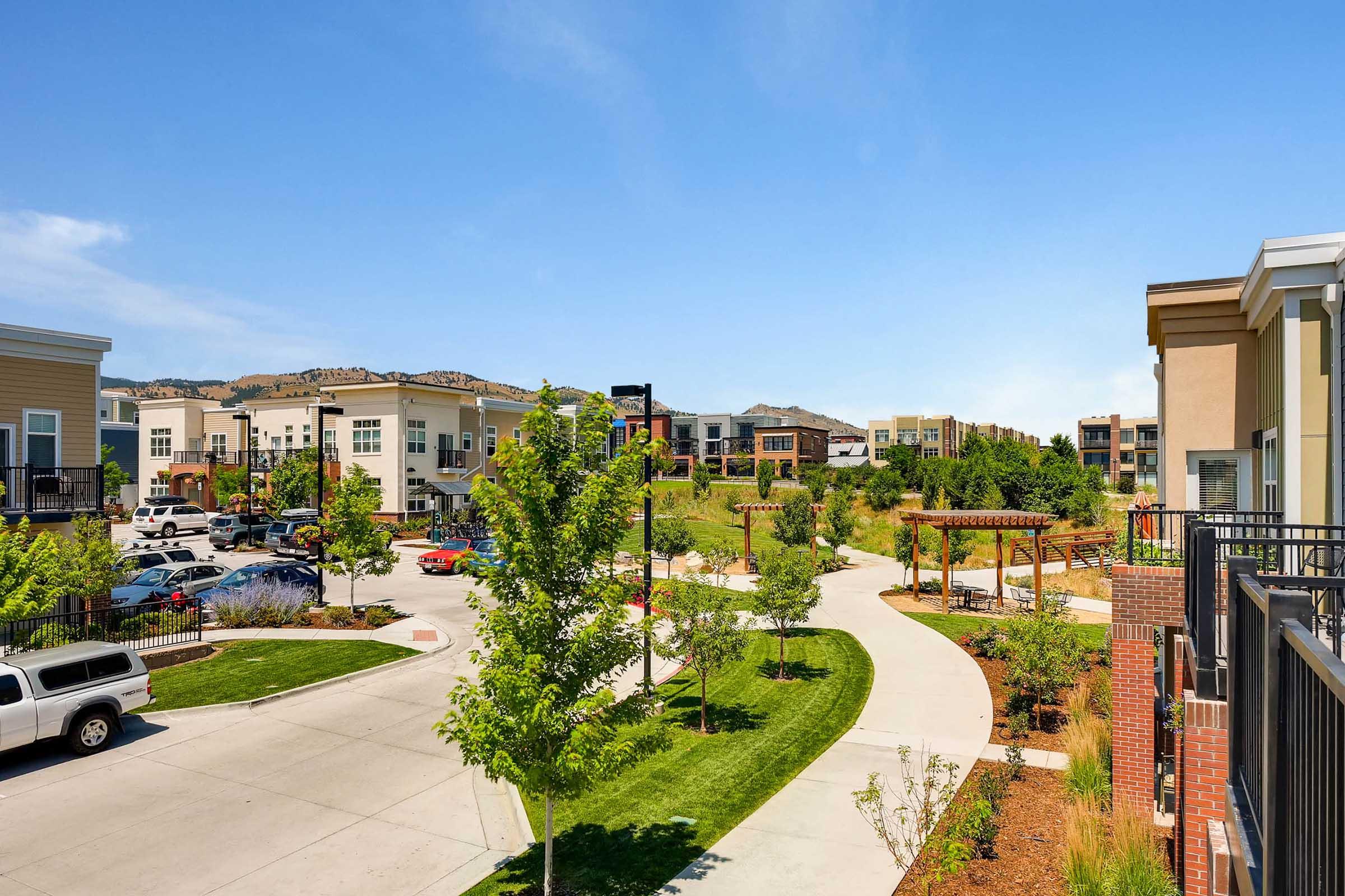 A modern residential neighborhood featuring multiple buildings with various architectural styles, green lawns, trees, and pathways. The scene includes parked cars and a pergola in the foreground, with mountains visible in the background under a clear blue sky.