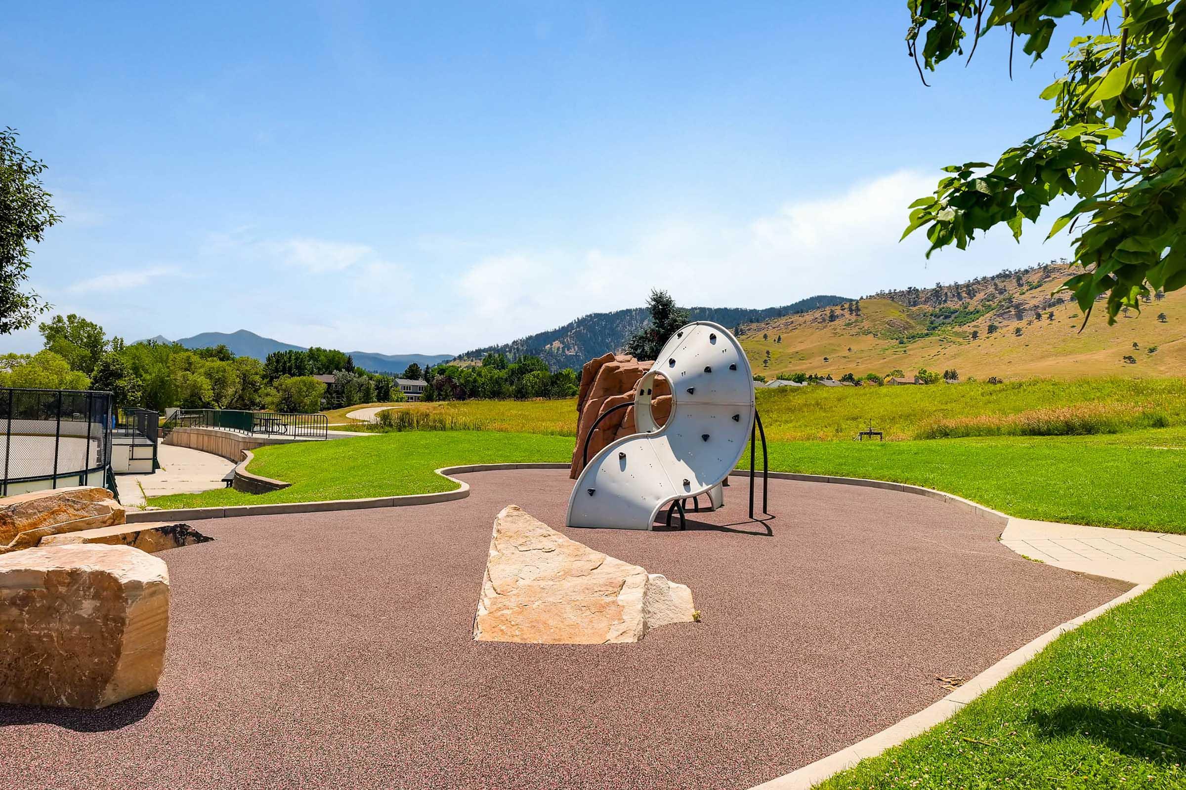 A playground with a climbing structure and large rock formations, set against a backdrop of rolling hills and blue sky. The ground is covered with rubber surfacing, and there is a pathway running through the grassy area. Trees flank the playground, creating a scenic outdoor space.