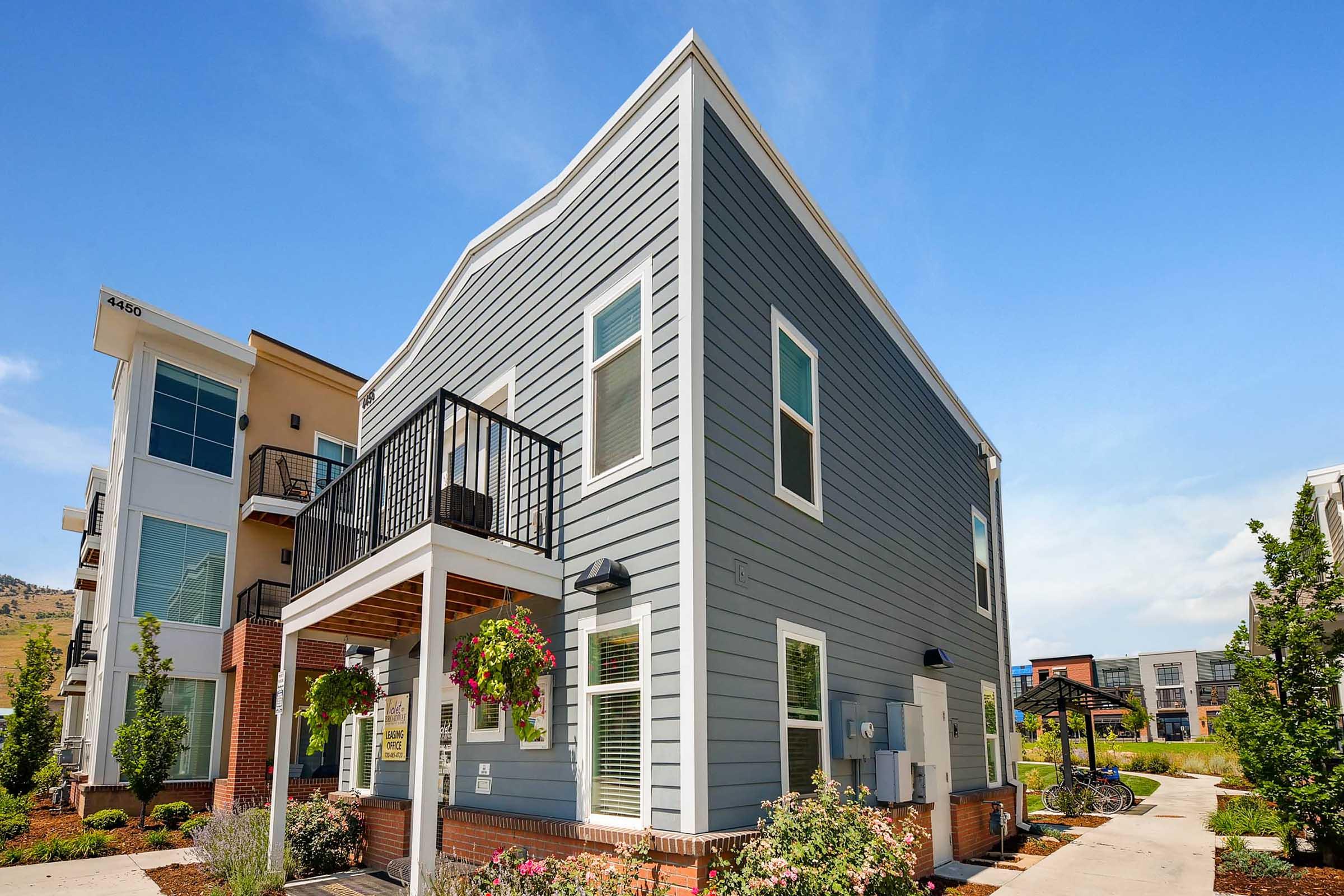 A modern gray townhouse with a balcony, surrounded by landscaped flower beds. Clear blue sky above and additional residential buildings in the background. The design features large windows and a clean, contemporary look, creating a welcoming atmosphere.