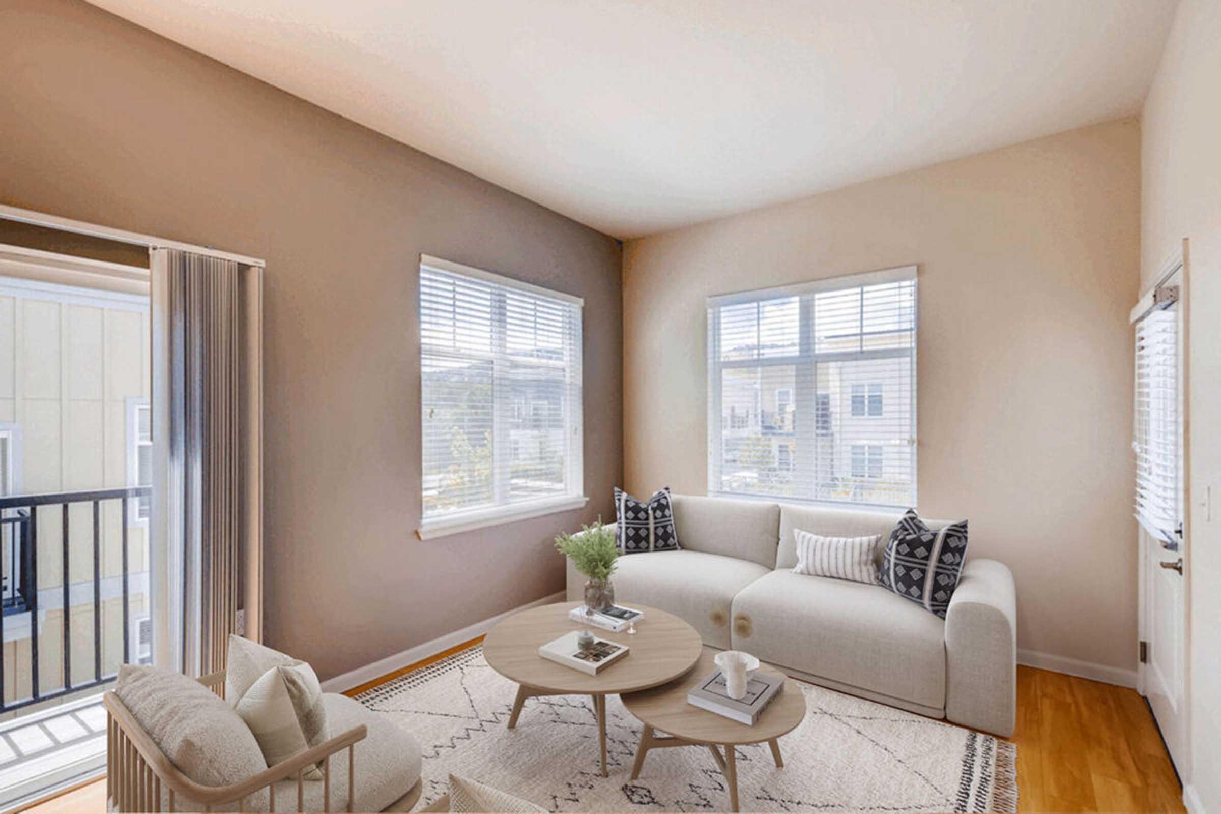 A bright living room featuring a light-colored sofa and an armchair, with a round coffee table in the center. Large windows allow natural light to fill the space, and a patterned area rug is laid on the wooden floor. A small plant decorates the table, adding a touch of greenery to the room.