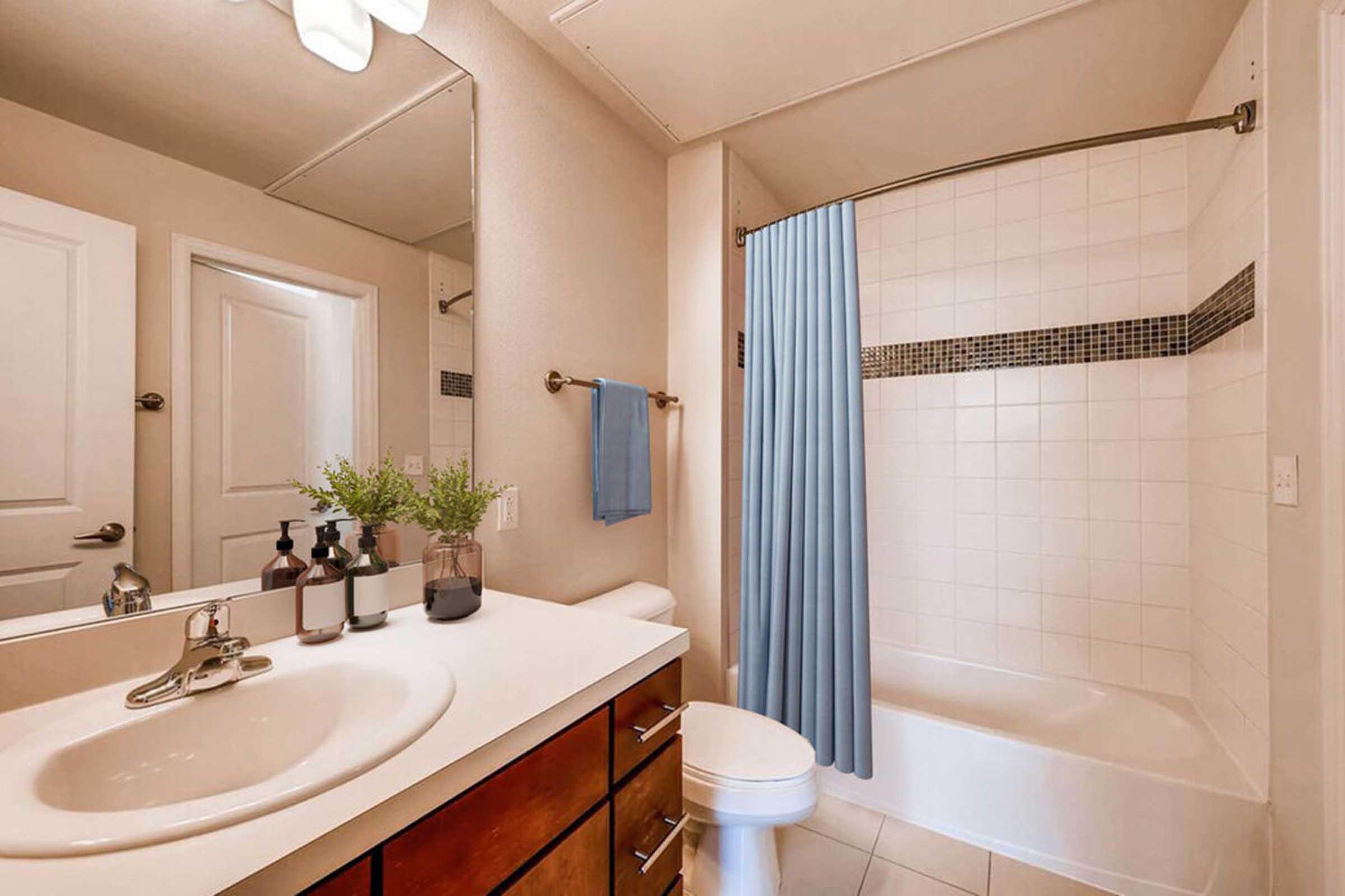 A modern bathroom featuring a white bathtub with a blue shower curtain, a sink with a countertop, decorative bottles, and a small plant. Light-colored walls and a towel hanging on a rack complete the clean and organized look. A door leads to another room, adding to the spacious feel.