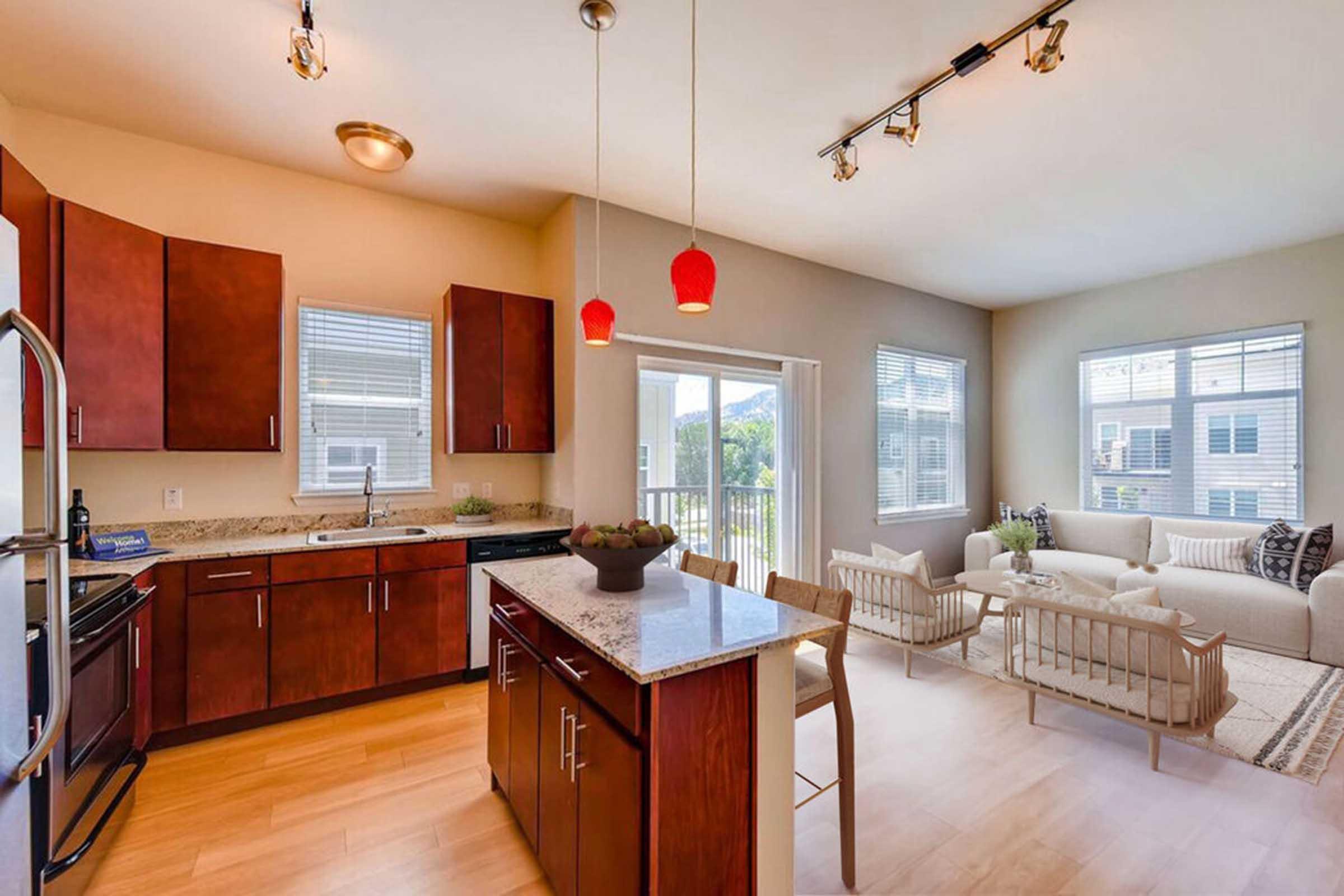 Modern kitchen with wooden cabinets, granite countertops, and stainless steel appliances. A small dining area features a central island with chairs. Natural light streams in through large windows, illuminating a cozy living space with a white sofa and decorative plants.