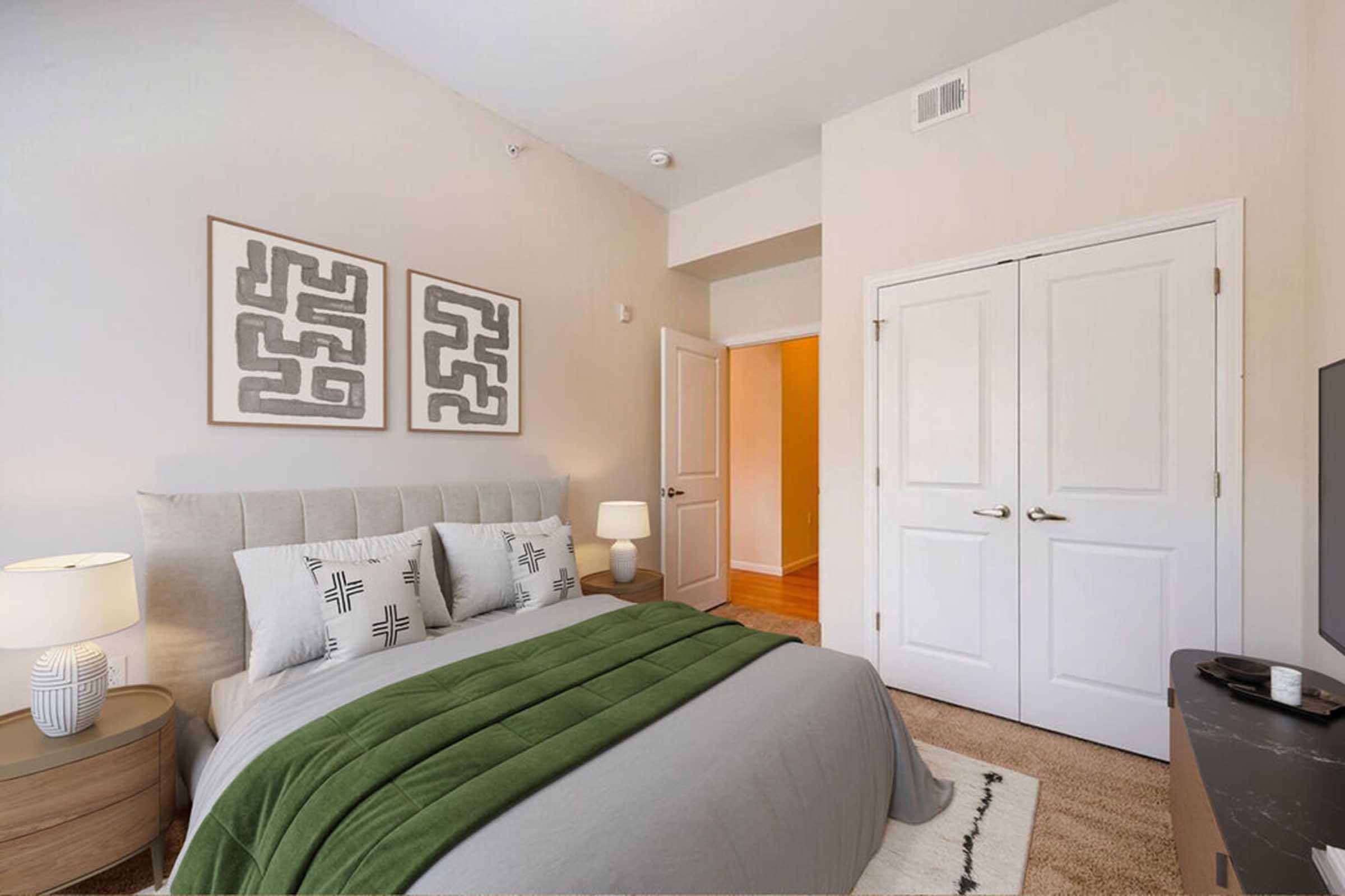 A neatly arranged bedroom featuring a light gray bedspread and decorative pillows. The room has two abstract wall art pieces, a bedside lamp on a round table, and a TV on a dresser. Two doors lead to other spaces, one of which is partially open, revealing a hallway. The overall color scheme is soft and inviting.