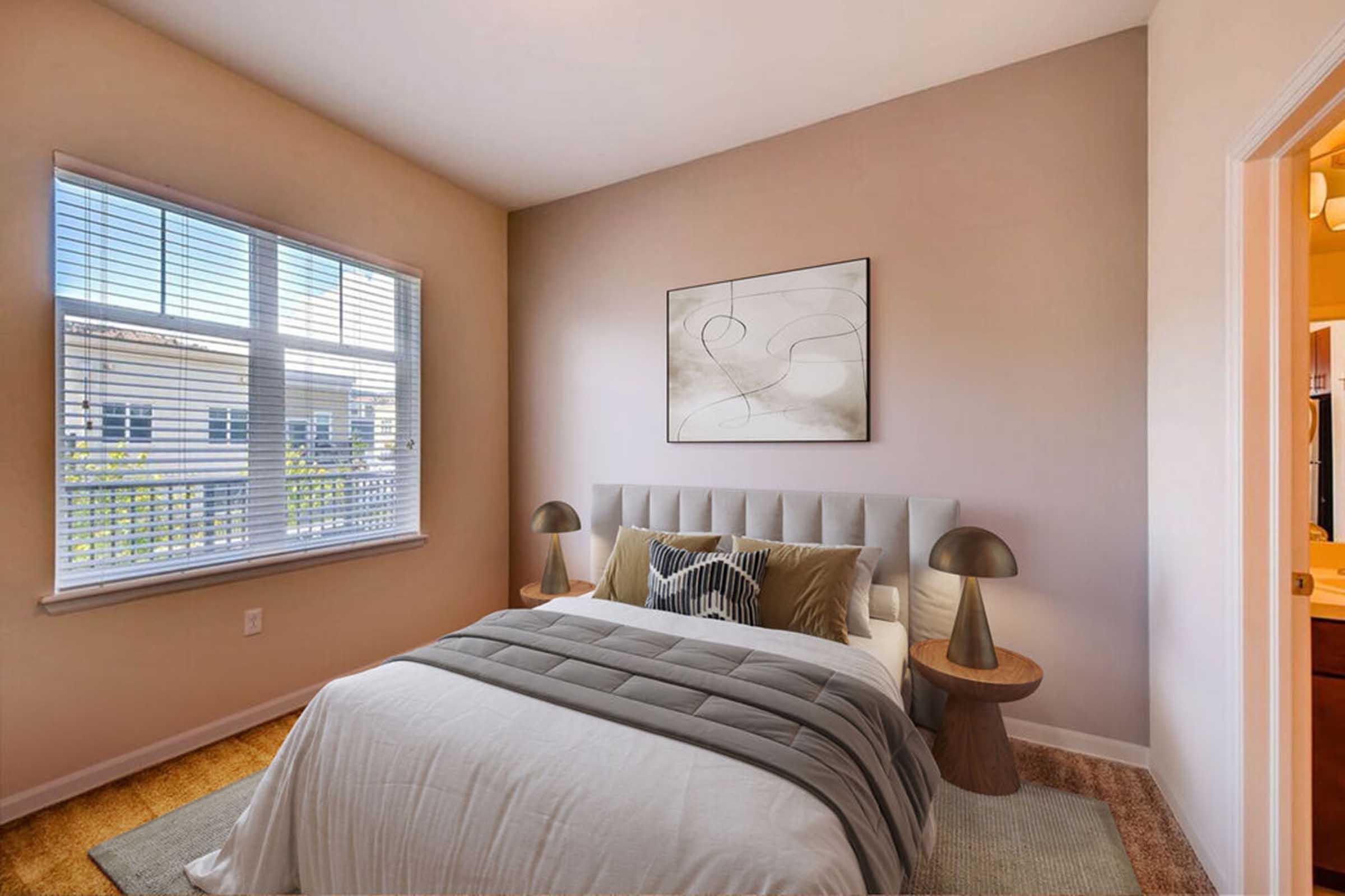 A cozy bedroom featuring a bed with a gray quilt and decorative pillows, flanked by two stylish lamps on wooden nightstands. A large window with blinds allows natural light to fill the room, and minimalistic artwork hangs on the wall, creating a serene atmosphere. Soft carpeting completes the inviting look.
