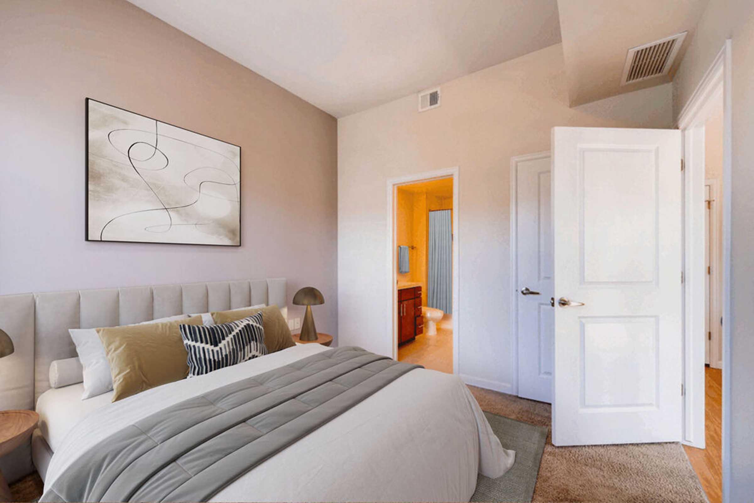 A well-decorated bedroom featuring a plush bed with neutral-colored bedding and decorative pillows. A large artwork hangs above the headboard. There are bedside lamps on each side of the bed, and a bright bathroom area is visible through an open door, showcasing modern fixtures and a blue shower curtain.