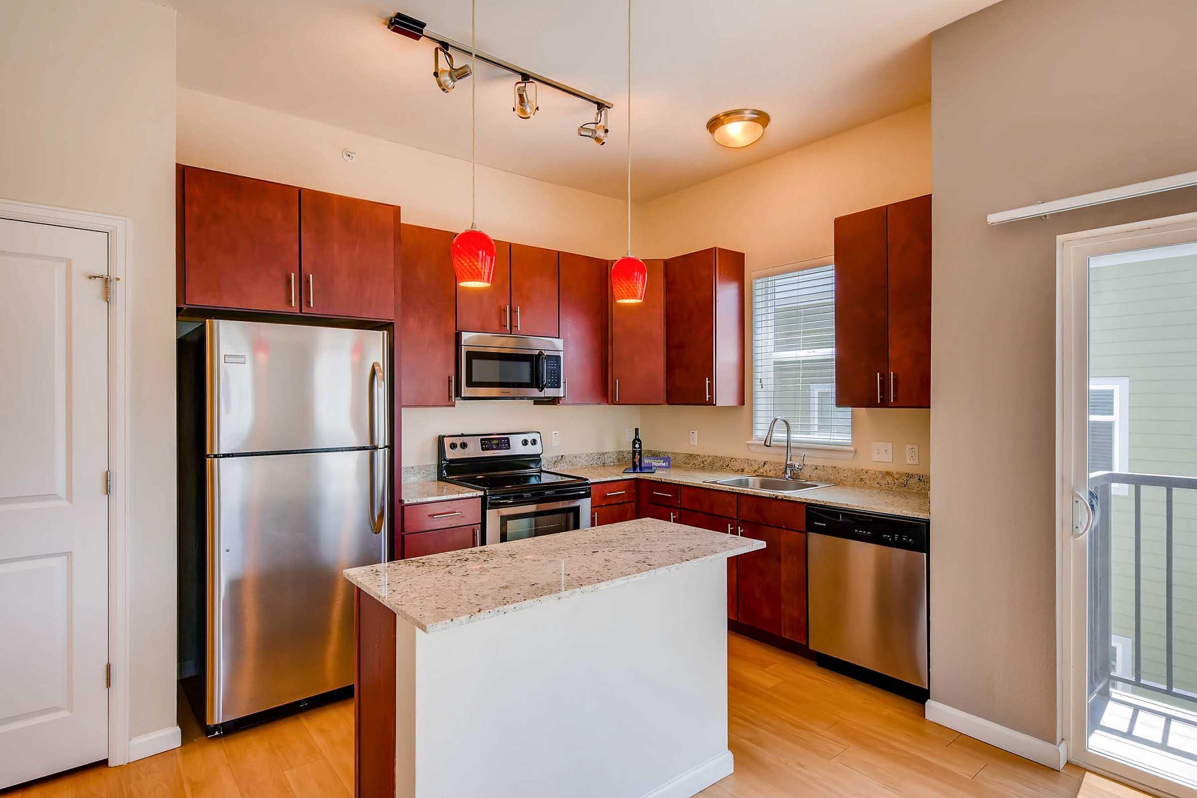 Modern kitchen featuring dark wood cabinets, stainless steel appliances including a refrigerator and oven, granite countertops, and pendant lighting. A large window lets in natural light, with a small balcony visible in the background. The floor is light-colored wood, adding warmth to the space.