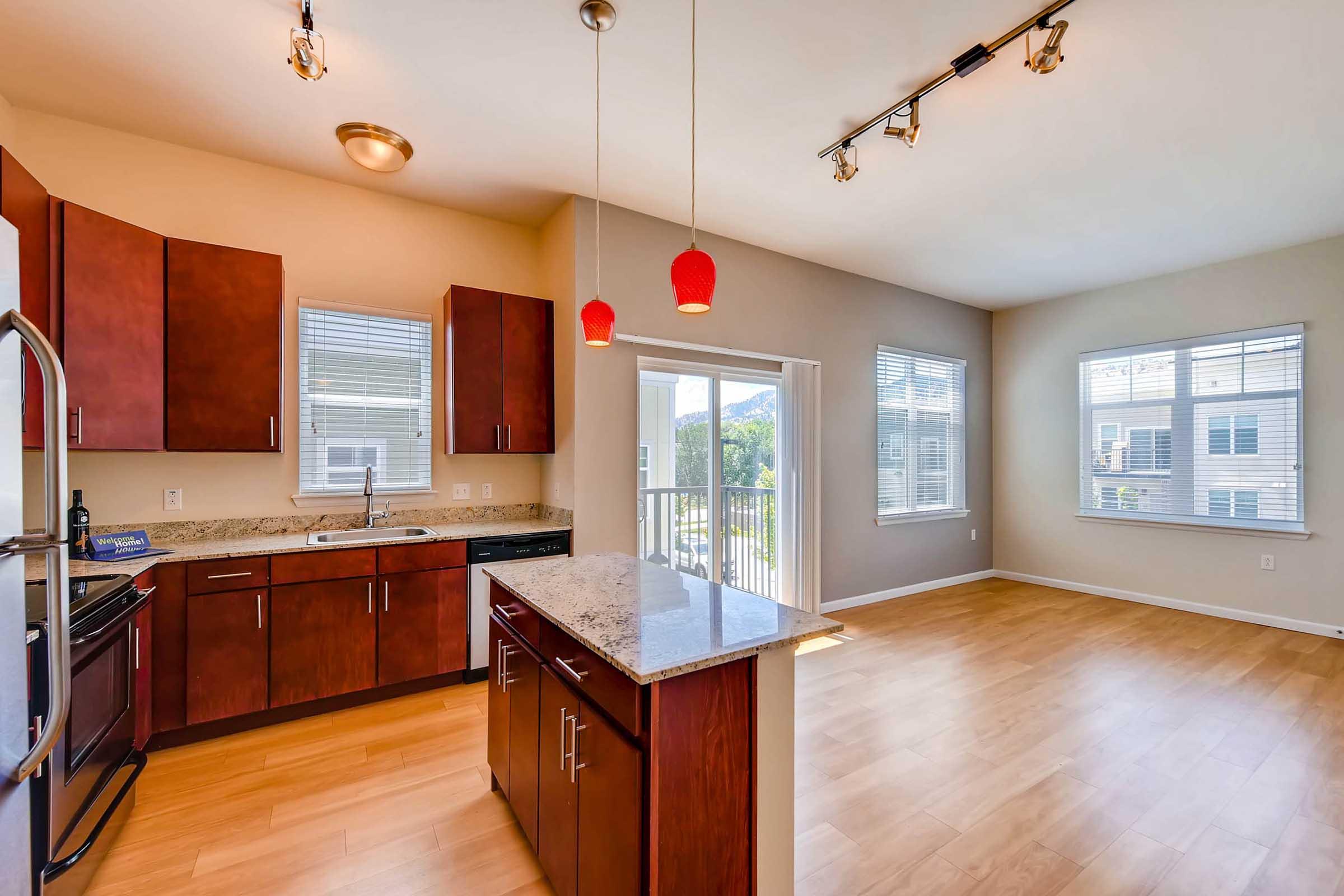 A modern kitchen and living area featuring dark wood cabinets, stainless steel appliances, and a granite countertop island. The space has large windows allowing natural light, with a view of the outdoors. Sleek pendant lights hang above the island, and the flooring is light wood.