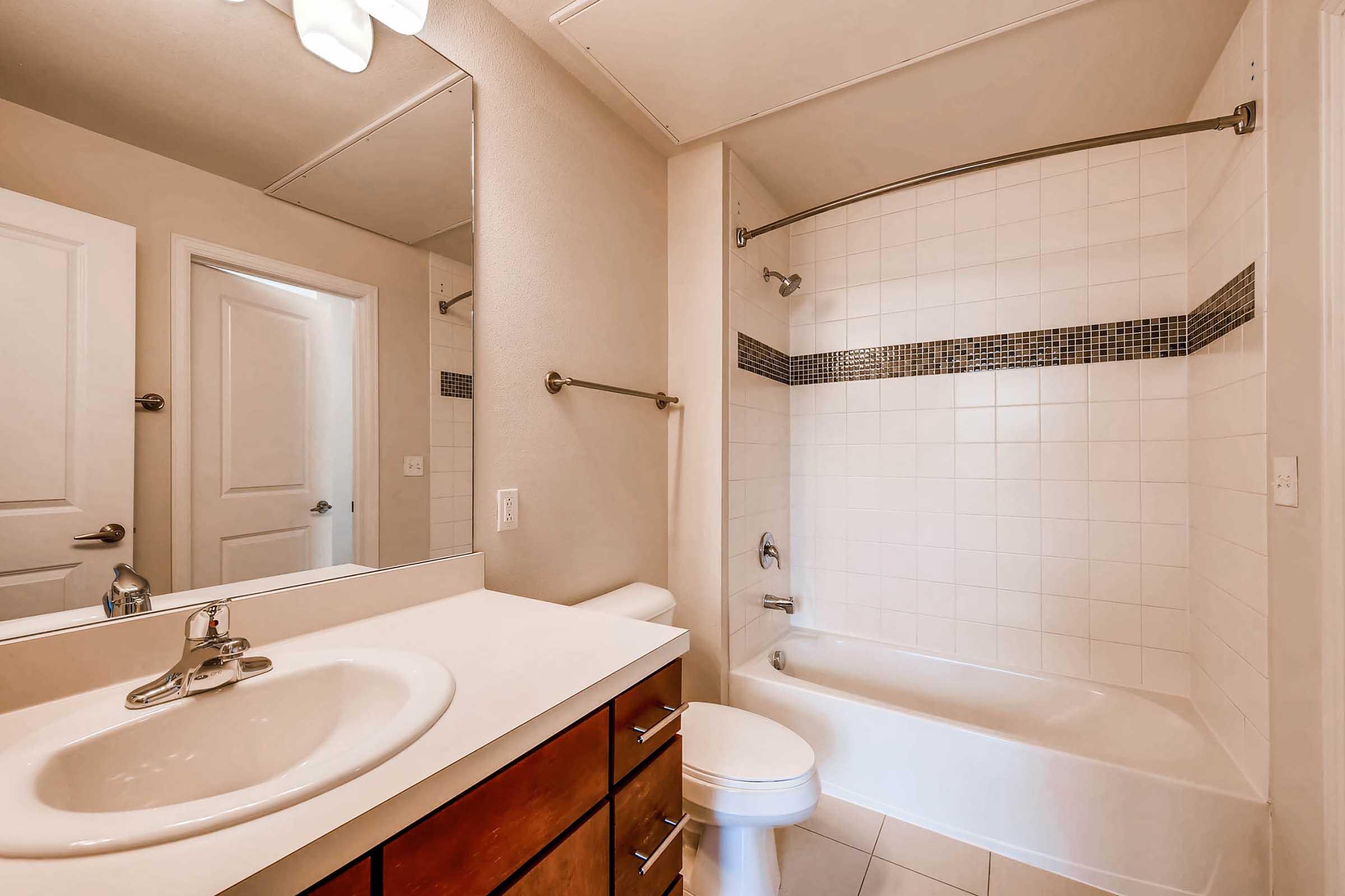 A clean bathroom featuring a white bathtub, a shower with a glass enclosure, and a large mirror above a sink with a simple countertop. The walls are tiled in light colors, and a light fixture is mounted on the ceiling. A door leads to another room in the background.