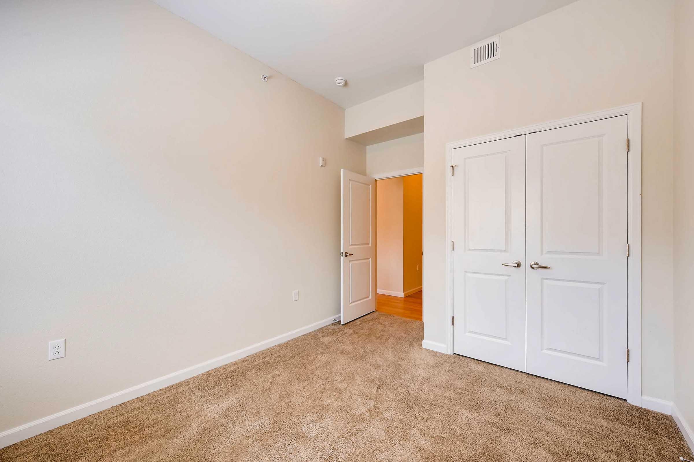 A light-colored room with beige carpet and cream walls. There are two double doors leading to a closet, and a doorway on the left provides access to another room. The space is empty and well-lit, creating a clean and inviting atmosphere.