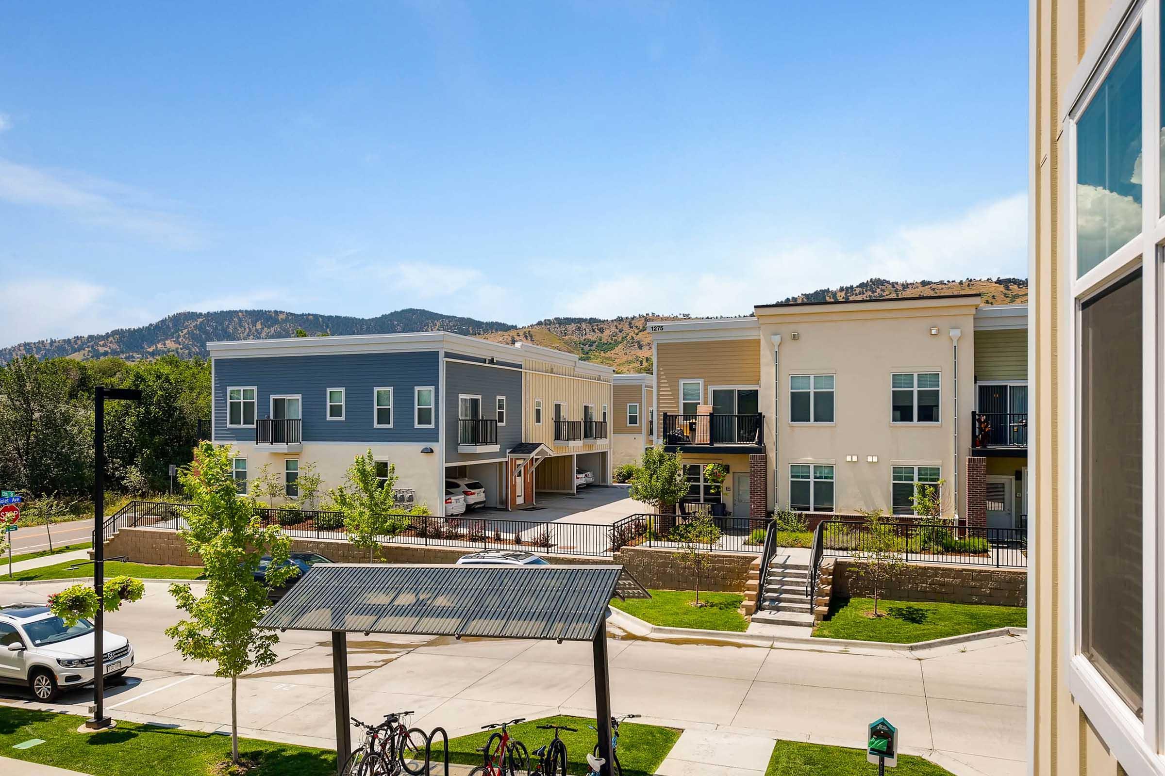 View of a modern residential complex with two-story buildings featuring balconies and landscaped greenery. In the foreground, there is a parking area, bicycles, and a covered seating structure. The background includes hills under a clear blue sky, creating a serene neighborhood atmosphere.