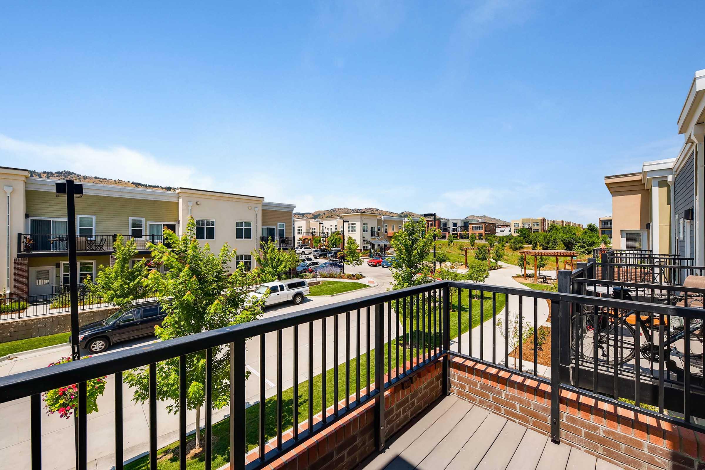 View from a balcony overlooking a landscaped residential area. The scene features multiple apartment buildings, greenery, and parked cars along a paved path. Bright blue sky enhances the vibrant atmosphere of the neighborhood.