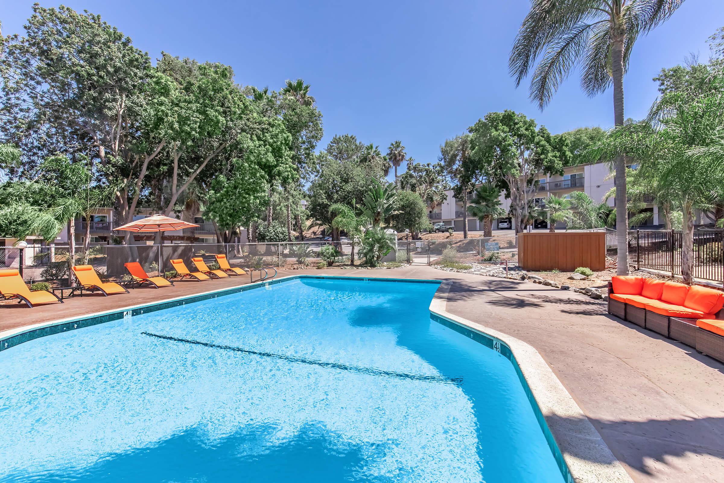 A vibrant outdoor swimming pool surrounded by lush greenery and palm trees. Bright orange lounge chairs line the pool area, while shaded umbrellas provide cover. The clear blue water reflects the sunny sky, creating a relaxing and inviting atmosphere. Adjacent areas feature pathways and seating arrangements.