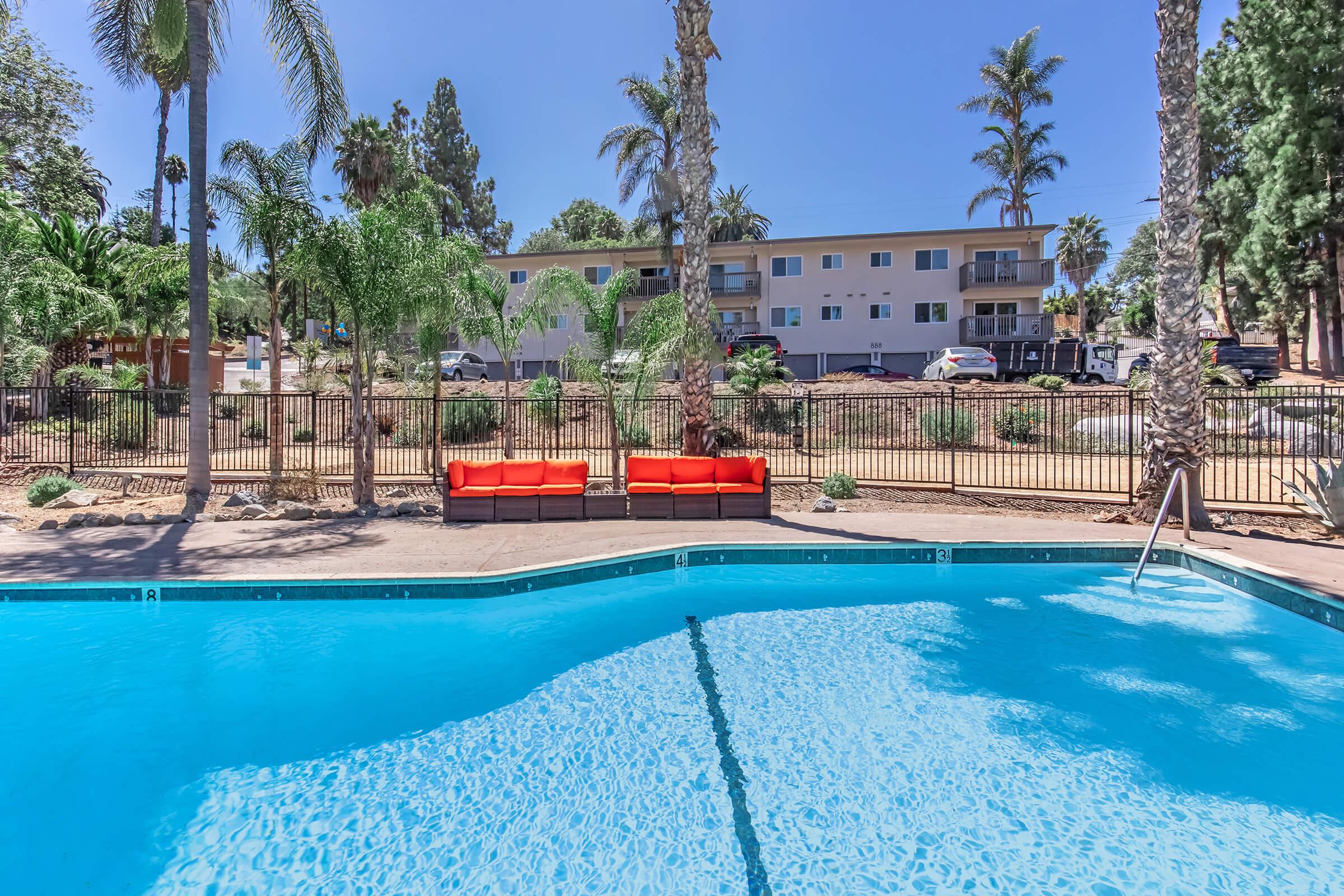 A sparkling blue swimming pool surrounded by palm trees, with an orange couch positioned beside it. In the background, a multi-story building is visible, set amidst a landscaped area with shrubs and grass under a clear blue sky.