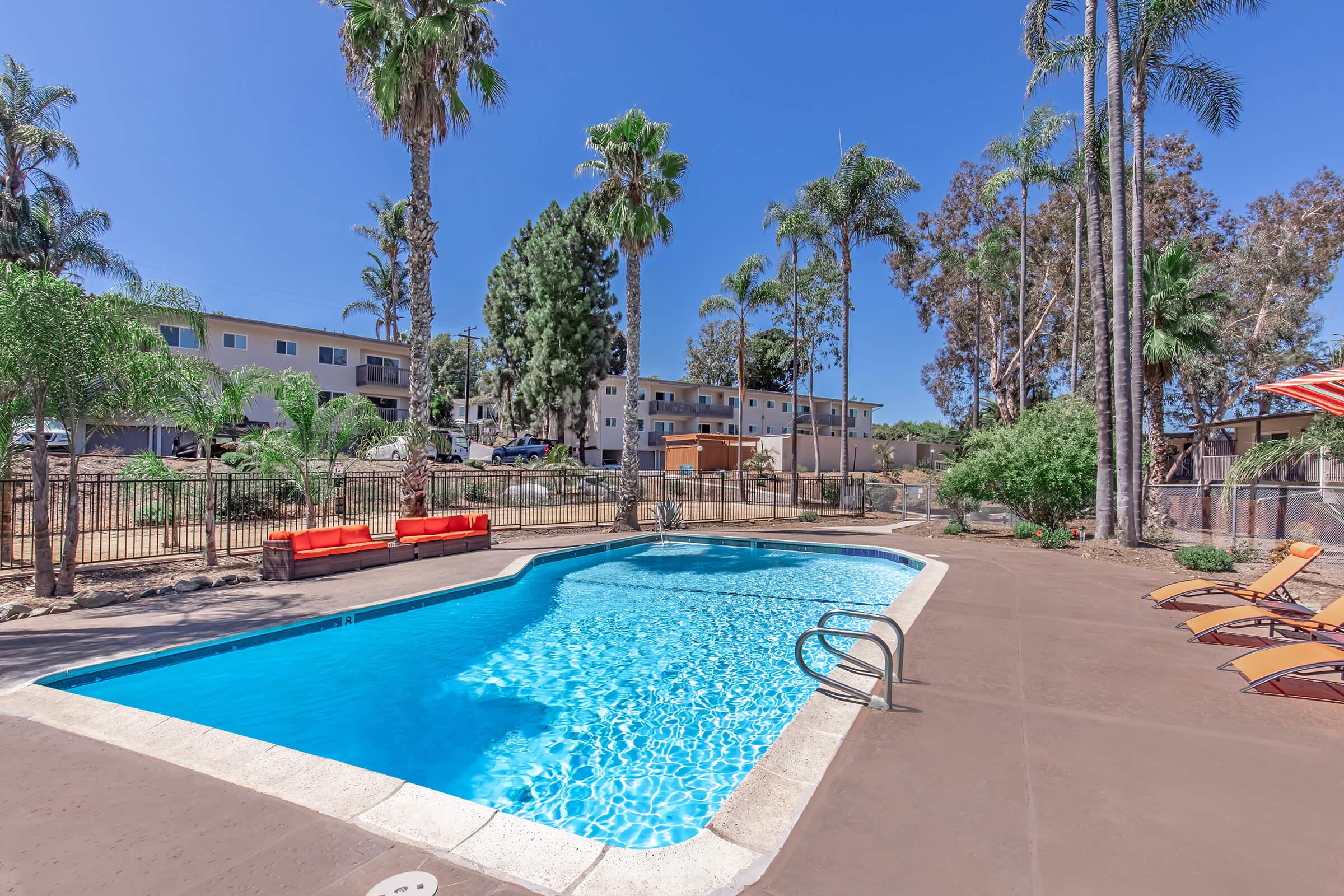 A serene outdoor swimming pool surrounded by palm trees and sun loungers, with vibrant red seating nearby. The clear blue water reflects the sunlight, creating a relaxing atmosphere. In the background, there are apartment buildings set among lush greenery, enhancing the tranquil setting.