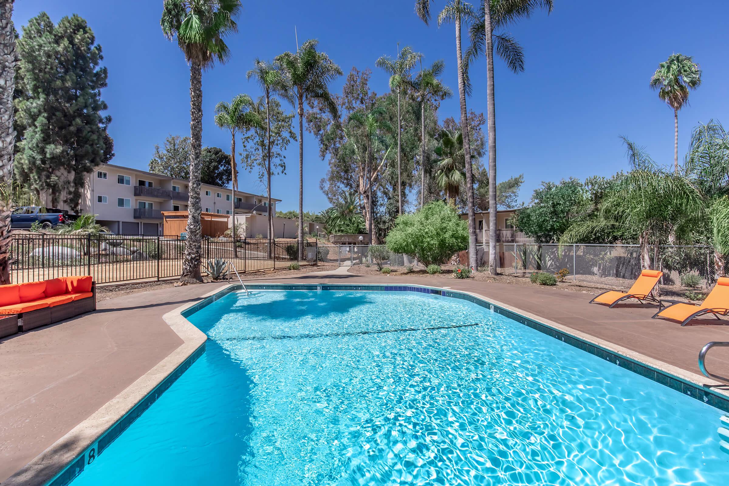 A bright outdoor swimming pool surrounded by palm trees, with orange lounge chairs on the side. The clear blue water reflects the sunlight. In the background, there are a few residential buildings and greenery, creating a relaxing and inviting atmosphere.