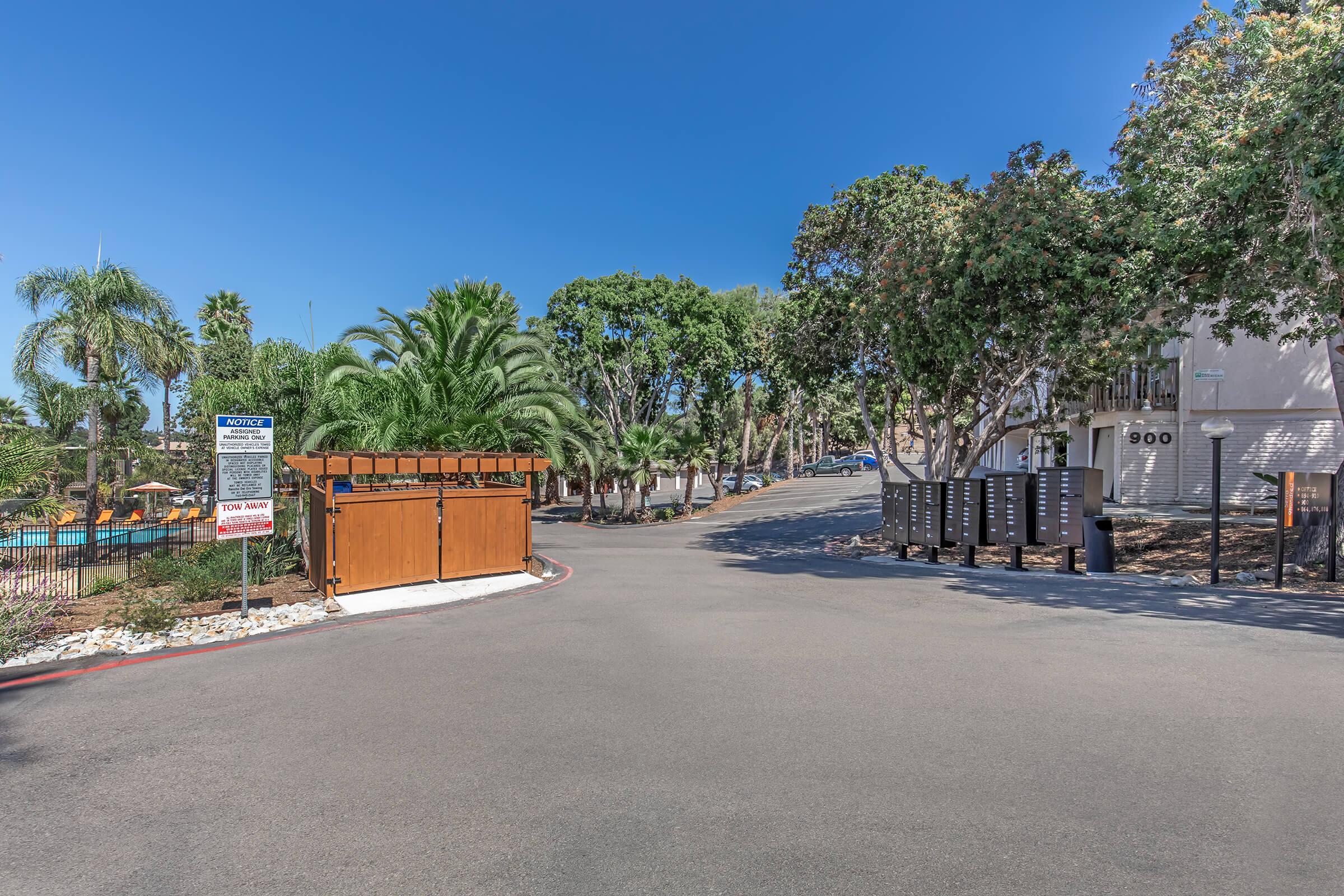A paved entrance to a residential area featuring palm trees and lush greenery. On the left, there is a wooden structure, possibly for trash or mail, and several mailbox units nearby. The clear blue sky provides a bright backdrop, with a swimming pool visible in the distance behind the gates.
