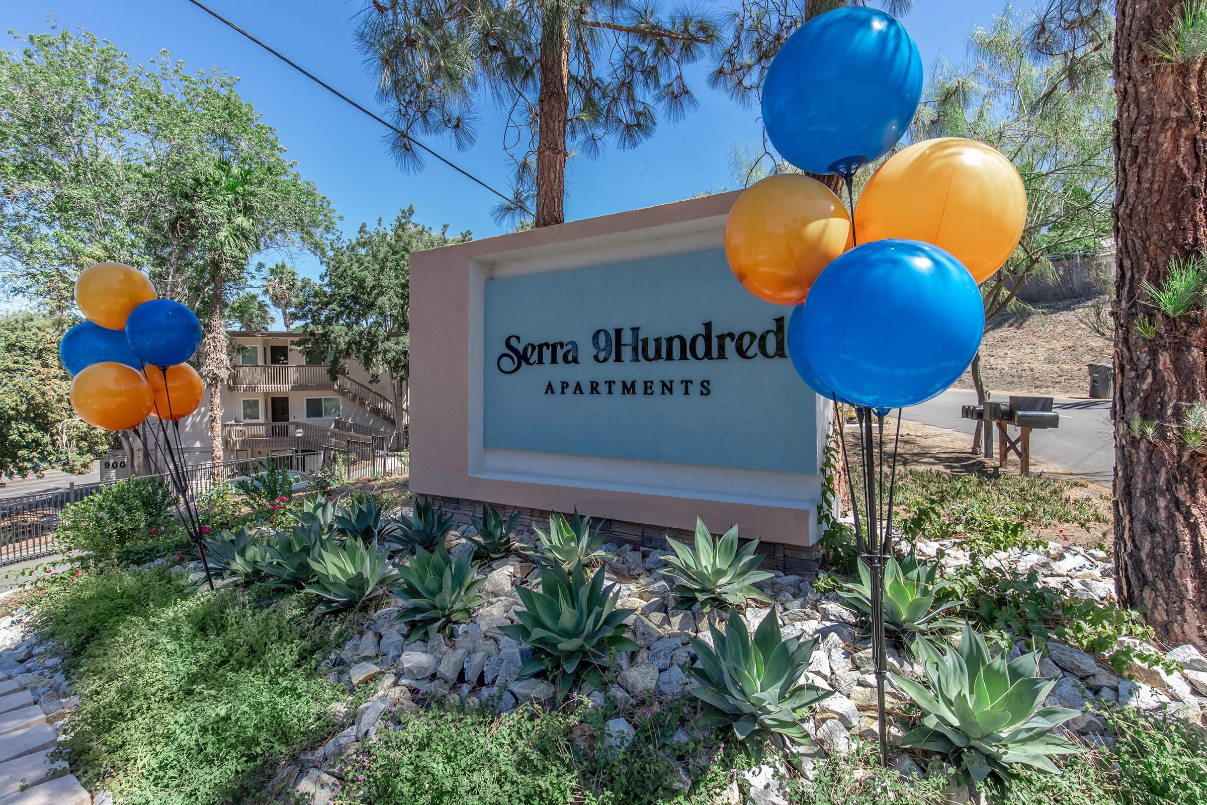 Sign displaying "Serra Hundred Apartments" surrounded by colorful blue and orange balloons. The area features landscaped greenery with rocks and succulents. Tall pine trees are visible in the background, and there are residential buildings on the hills behind the sign. Clear blue sky overhead.