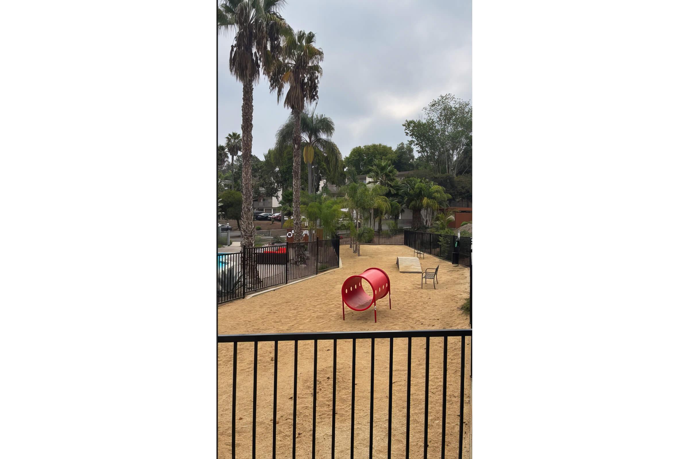 A playground area with sandy ground, featuring a red tube slide, a bench, and several palm trees. In the background, there are glimpses of a pool and surrounding foliage, with a cloudy sky overhead. The area is enclosed by a black fence.