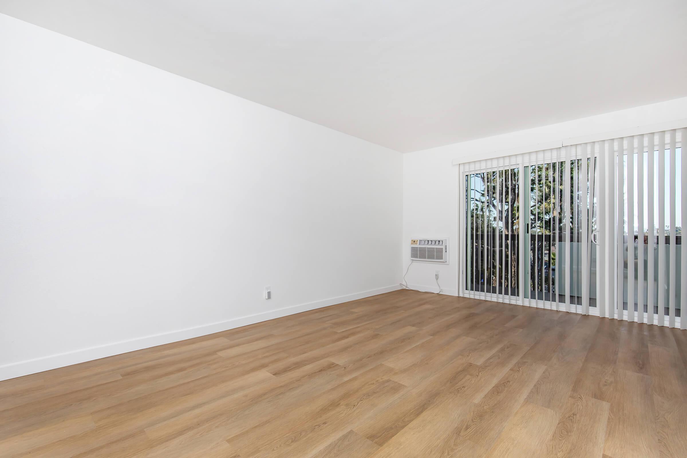 Empty room with light wood flooring and white walls. There is a large window with vertical blinds letting in natural light. An air conditioning unit is visible on the wall to the left. The space is uncluttered and minimalistic, creating a sense of openness.