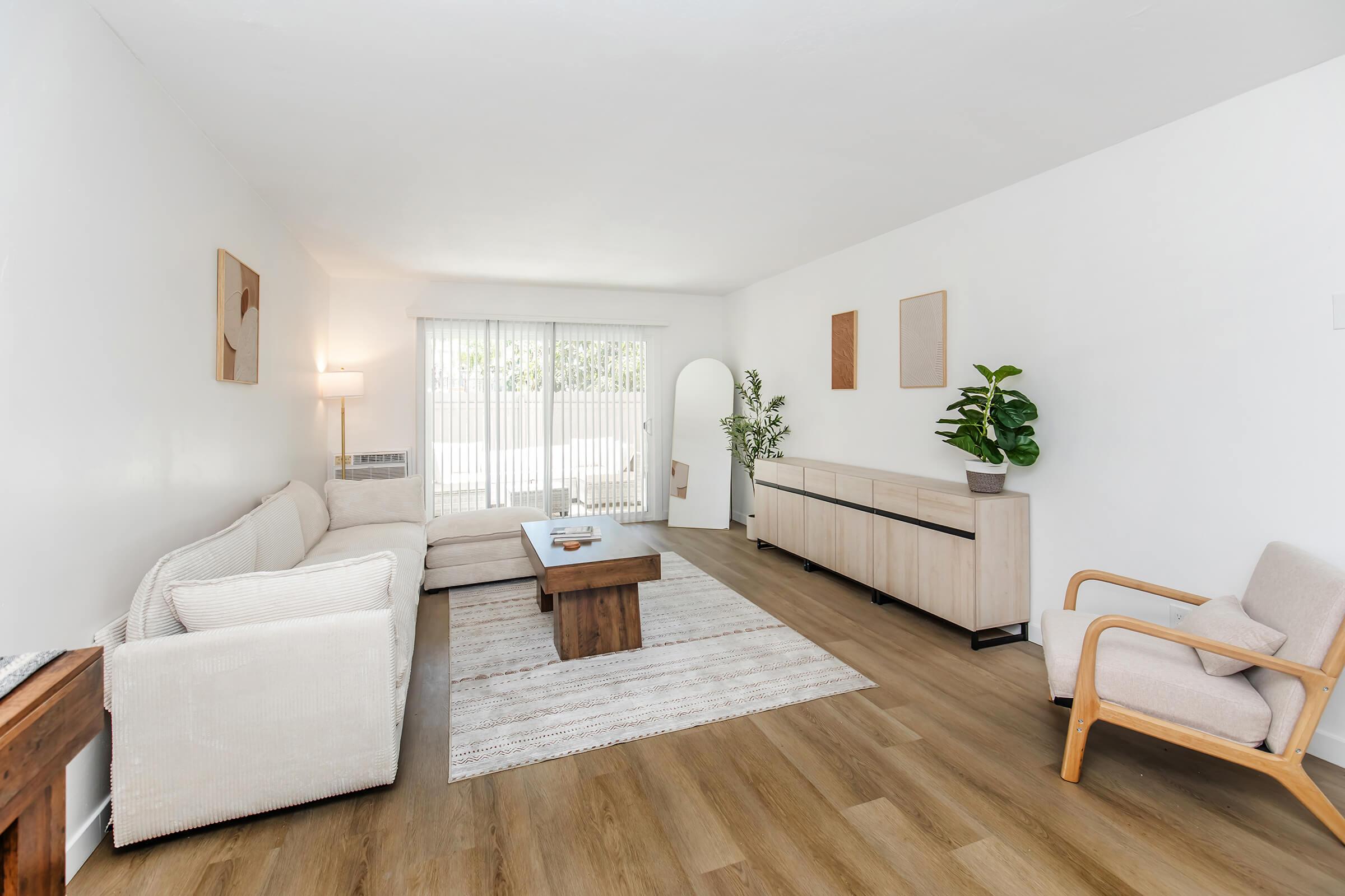 A bright, modern living room with a light color scheme. There’s a beige sofa and a wooden coffee table in the center, with a small plant on the table. A wooden sideboard against the wall complements the decor. Sunlight filters through sheer curtains, creating a warm atmosphere. A relaxing, minimalist aesthetic is present.