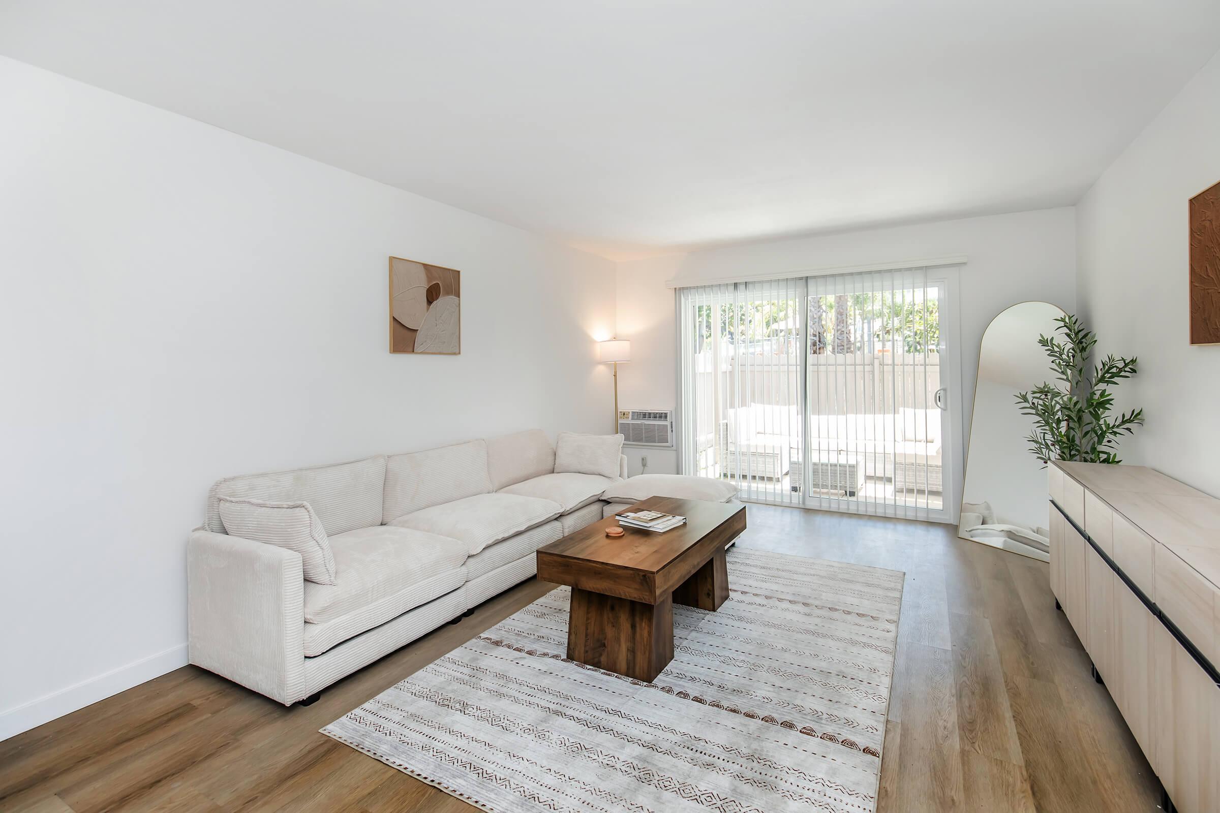 A bright and airy living room featuring a soft beige sofa, a wooden coffee table, and a light-colored area rug. Large glass doors provide access to an outdoor space, and a modern floor lamp stands in the corner. Decorative wall art adds a touch of style, while potted greenery enhances the inviting atmosphere.