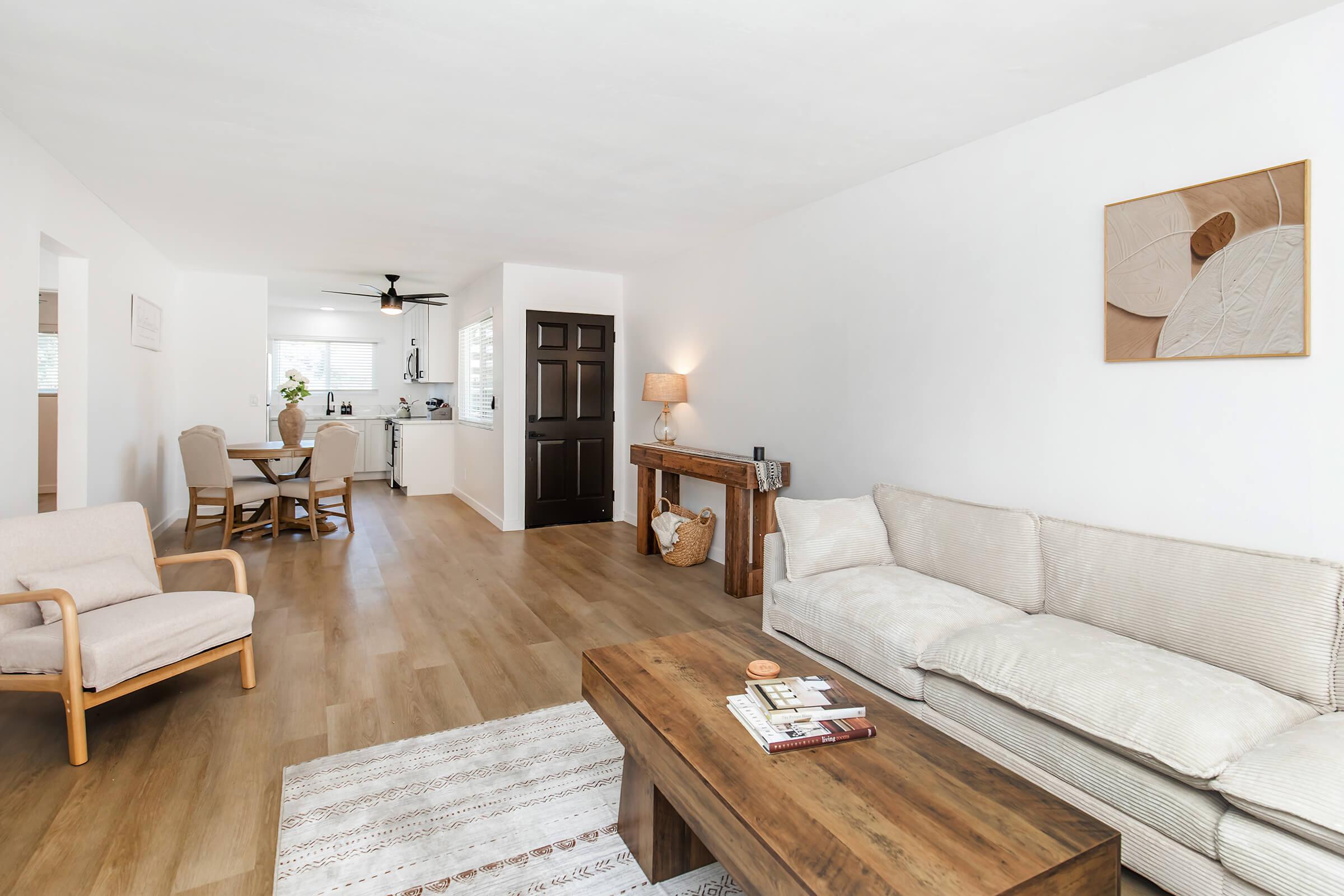 A bright, modern living room featuring a beige sofa, a wooden coffee table with magazines, and a light rug. In the background, there's a dining area with a table and chairs. The walls are white, and a small lamp provides soft lighting, creating a warm and inviting atmosphere.