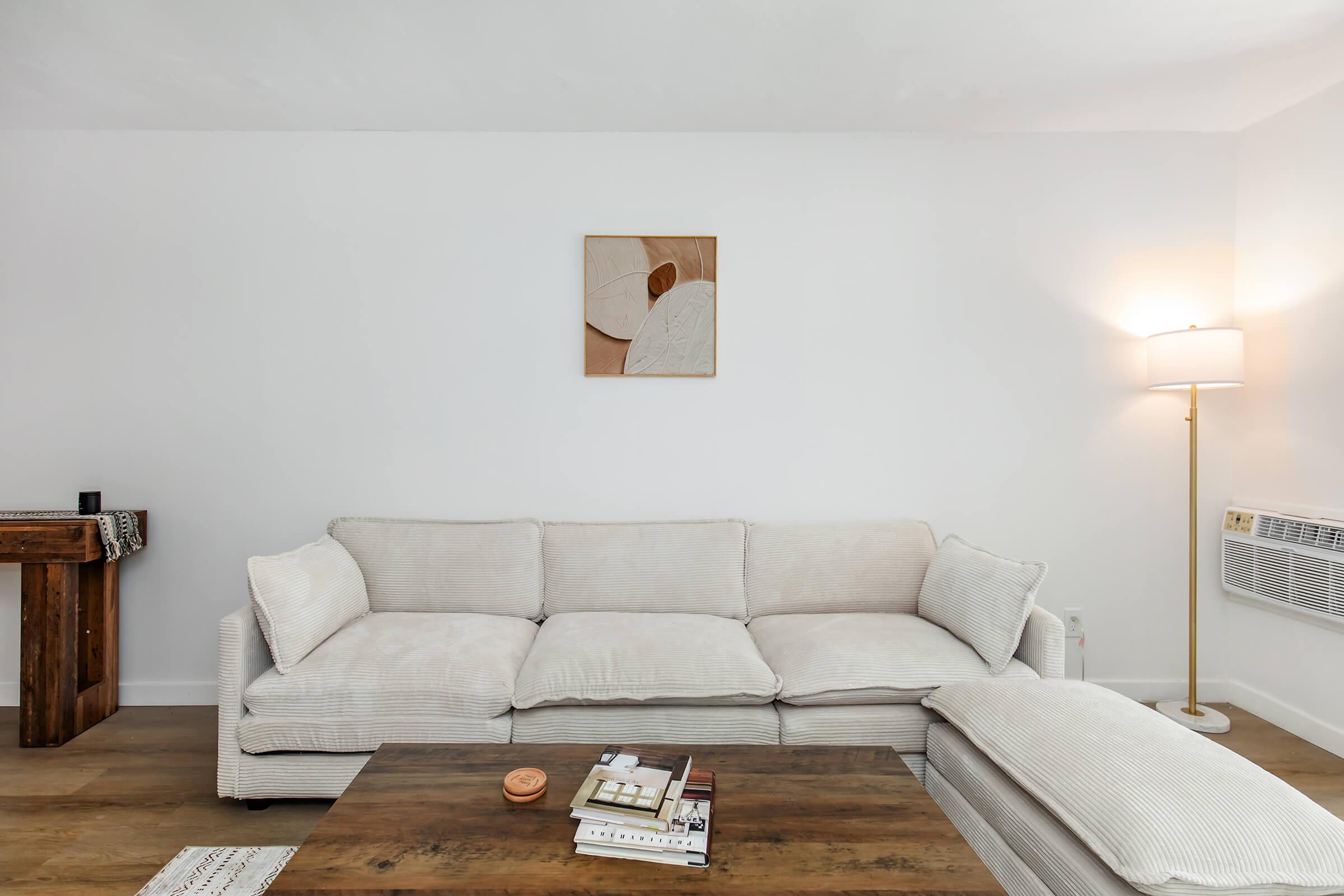 A cozy living room featuring a light-colored sectional sofa, a wooden coffee table with books and a candle, and a side table with a lamp. The walls are white, and there is a minimalist artwork hanging above the sofa, contributing to a warm and inviting atmosphere.