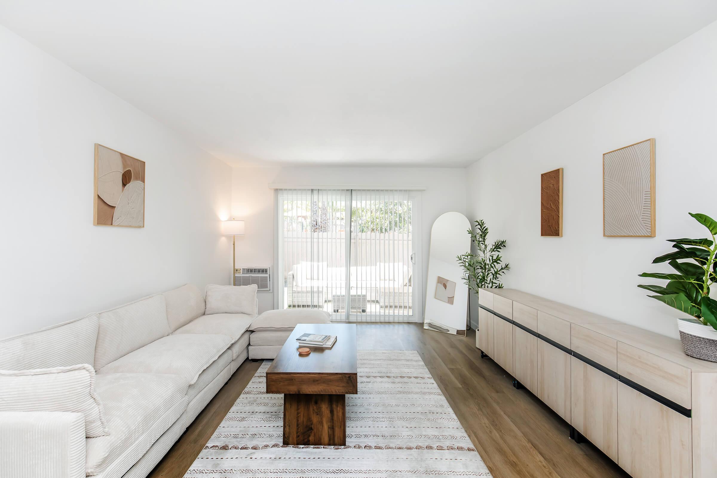 Modern living room featuring a light-colored sectional sofa and a wooden coffee table. The decor includes abstract wall art, a floor lamp, and a decorative plant. A sliding glass door leads to an outdoor area, allowing natural light to fill the space, with neutral tones and minimalistic design throughout.