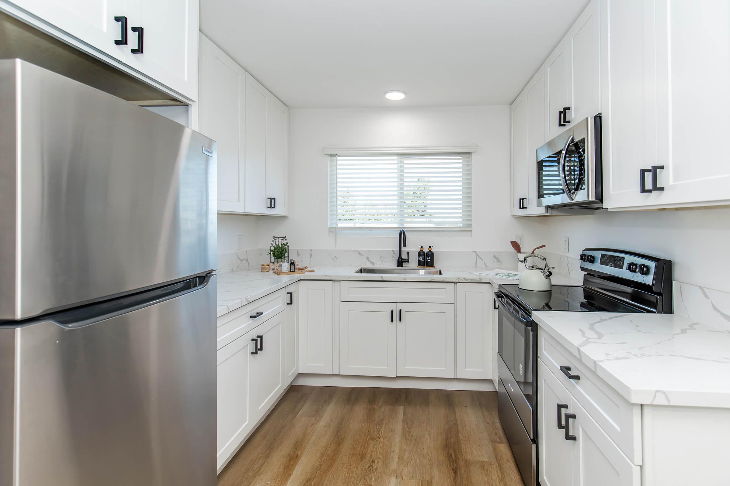 Modern kitchen featuring white cabinetry, stainless steel appliances, and marble countertops. A large fridge is on the left, with an oven and microwave on the right. The sink is placed below a window with blinds, allowing natural light. Wood flooring completes the contemporary look.