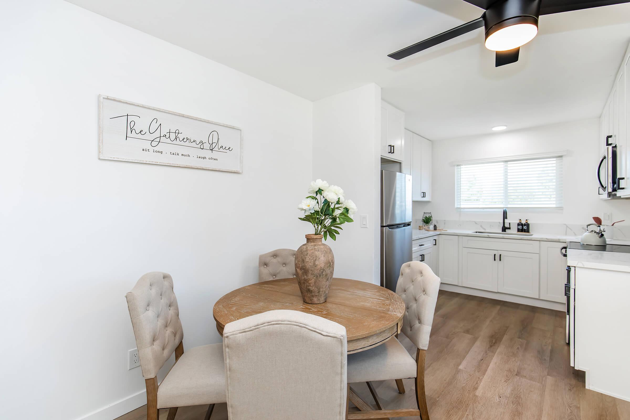 A bright and modern kitchen with a dining area featuring a round wooden table and two upholstered chairs. A decorative vase with white flowers sits on the table. The kitchen has white cabinetry, a window letting in natural light, and a minimalist sign on the wall that reads "The Gathering Place."