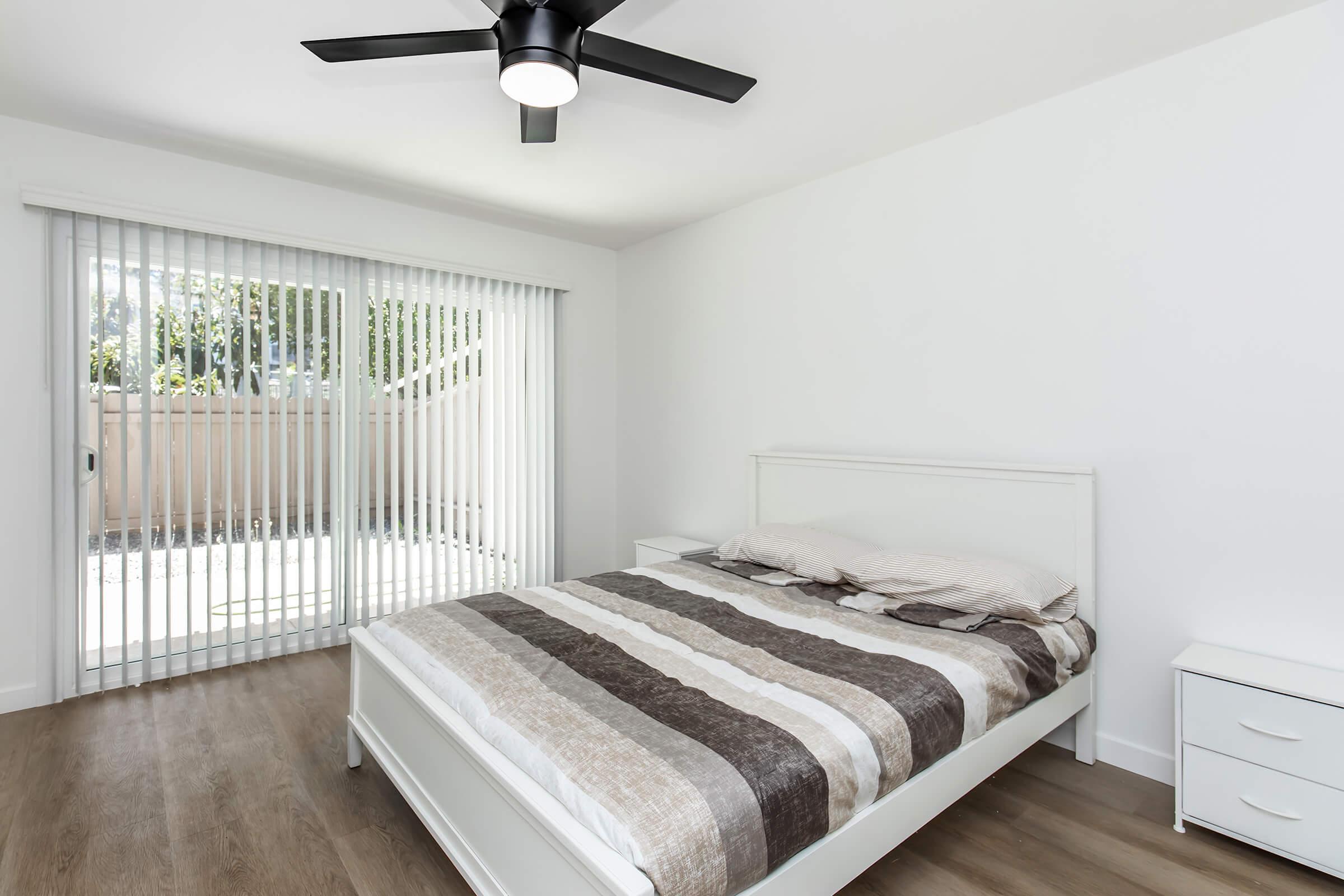 A bright and modern bedroom featuring a white bed with a striped comforter, a nightstand, and a ceiling fan. There is a large window with vertical blinds that allows natural light to enter, revealing an outdoor patio area. The flooring is light wood, adding to the room's airy feel.