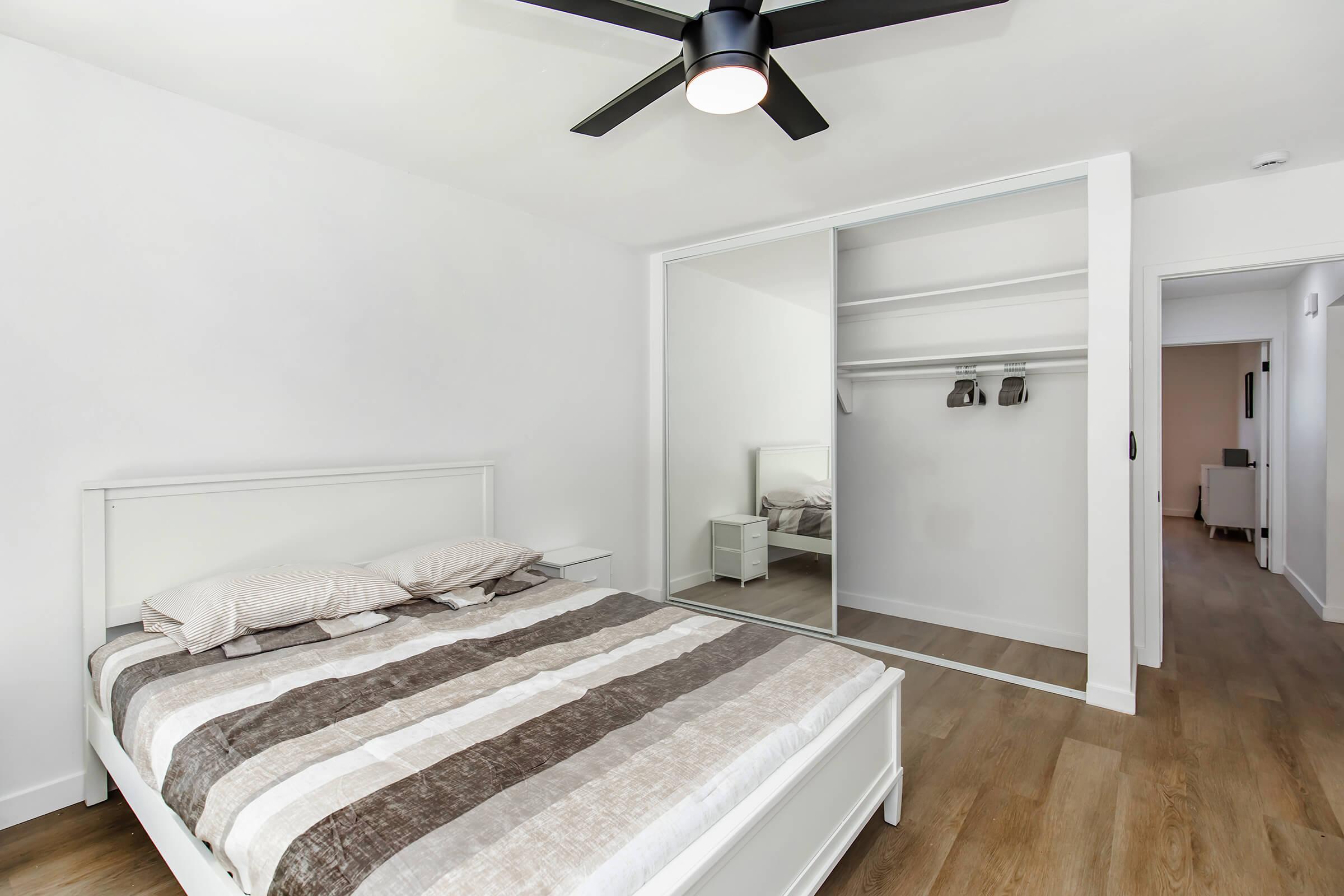 A modern bedroom featuring a white bed with a striped comforter, a ceiling fan, and a mirrored closet. The room has light wood flooring and minimalistic decor, creating a spacious and airy atmosphere. A doorway leads to another room in the background.