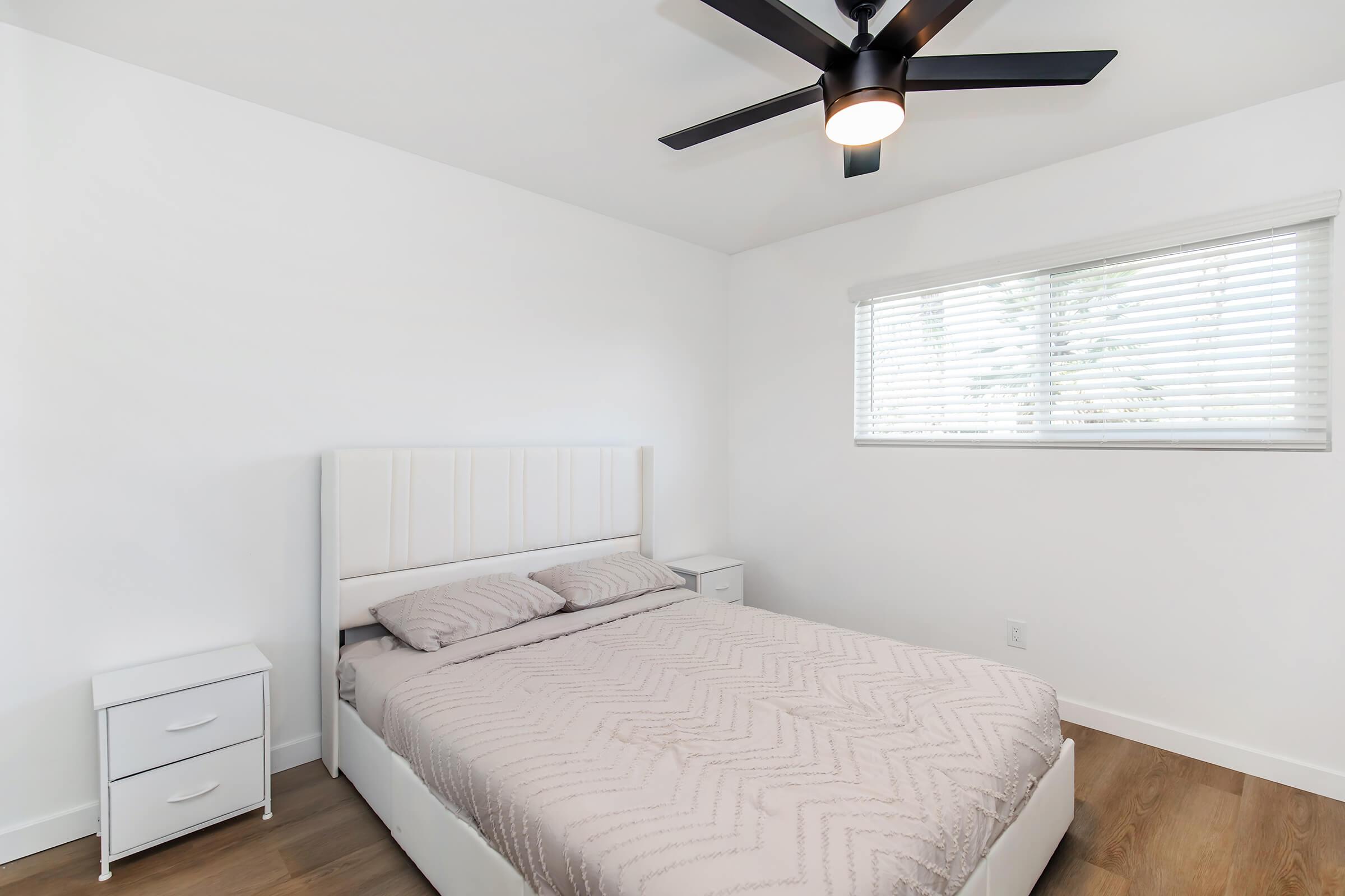 A clean, modern bedroom featuring a light-colored bed with a patterned duvet, a nightstand on either side, and a ceiling fan above. There is a window with blinds allowing natural light to enter, illuminating the minimalist decor in the room.