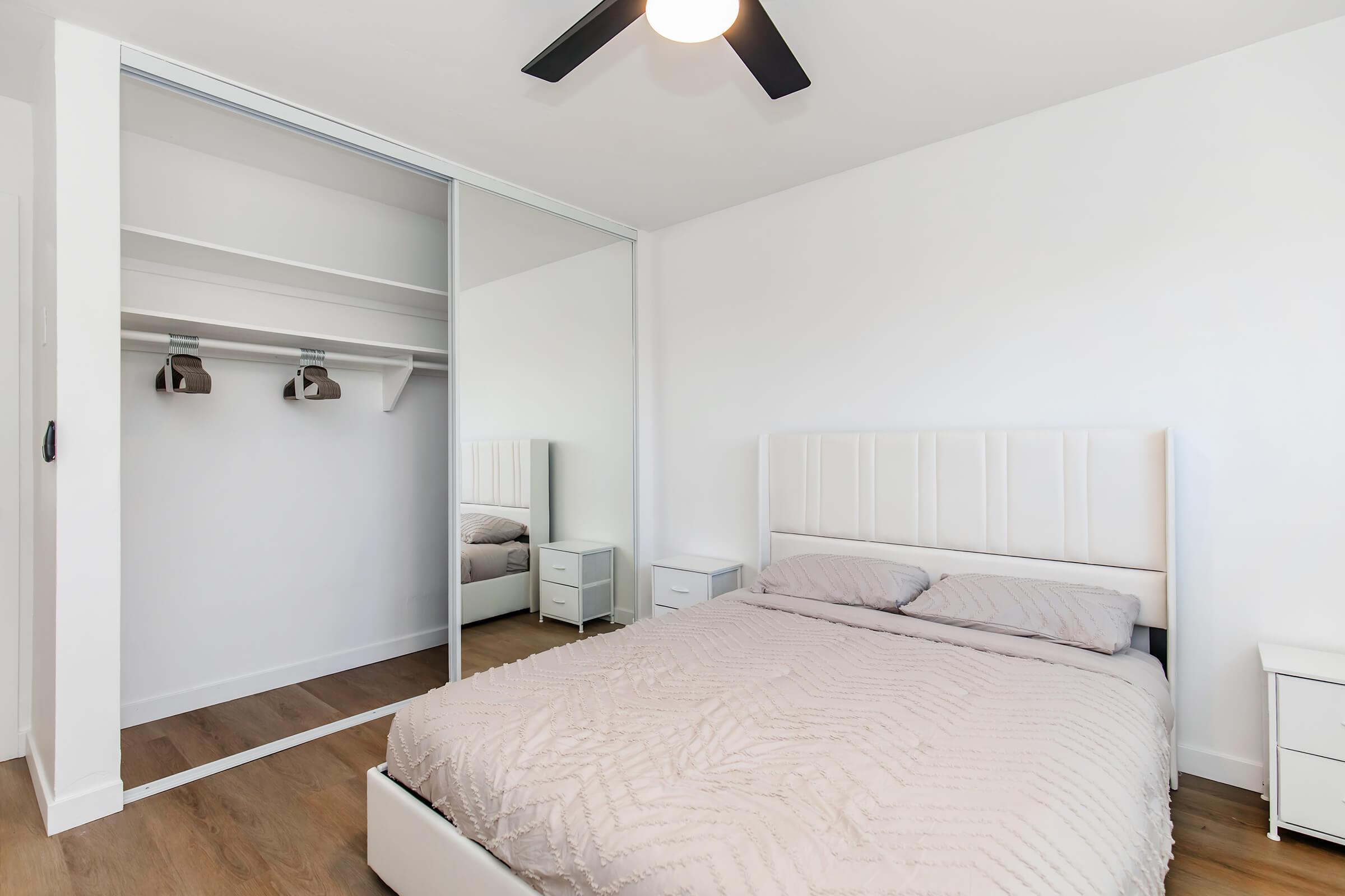 A modern bedroom featuring a white bed with a textured bedspread, two small nightstands on either side, and a large mirrored closet reflecting the room. The walls are white, and there is a ceiling fan above, creating a bright and spacious atmosphere.