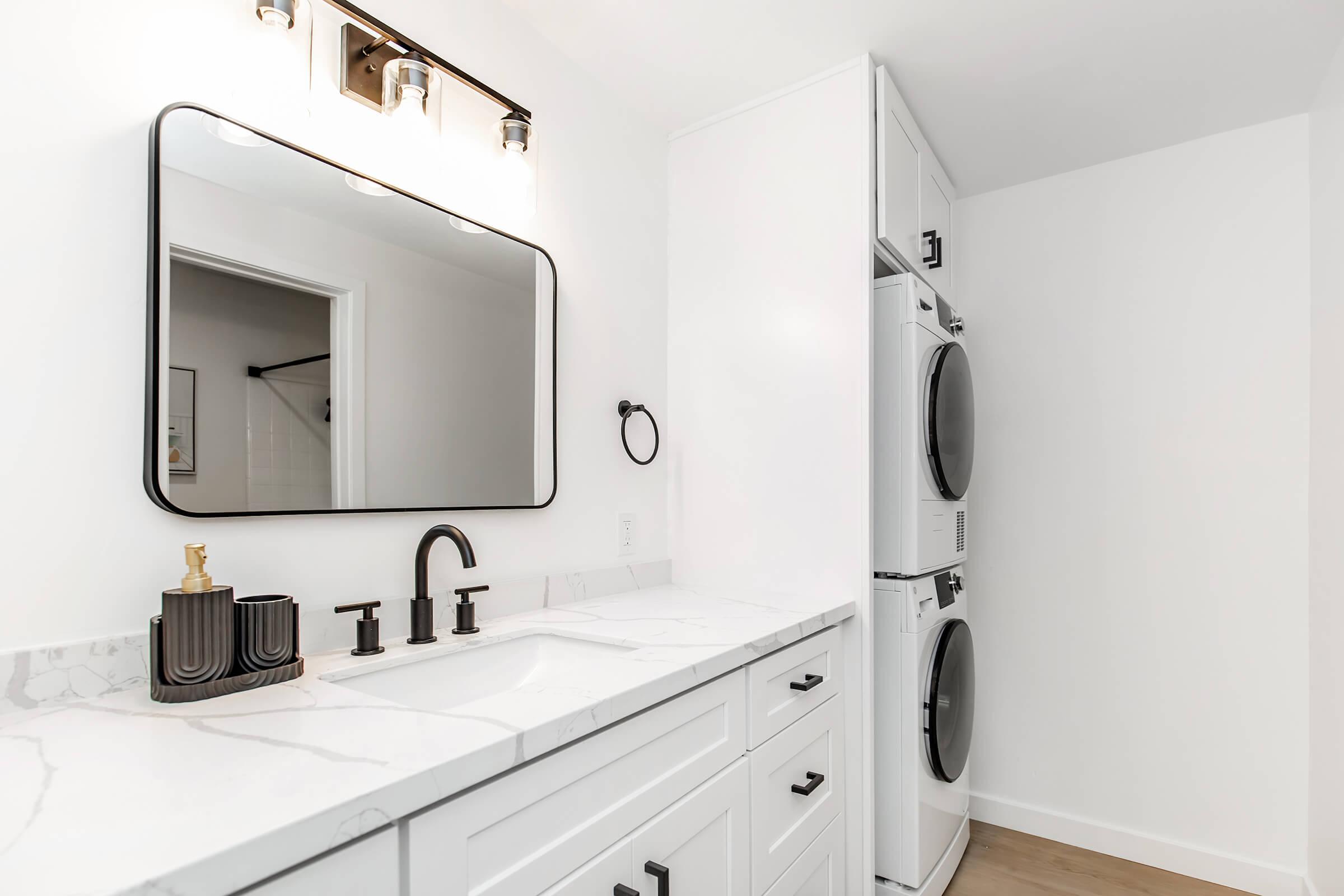 A modern laundry room featuring a stacked washer and dryer in a white cabinet, a sleek marble countertop with a black faucet and sink, a round mirror above the sink, and minimalistic decor. The walls are painted white, contributing to a clean and bright aesthetic.