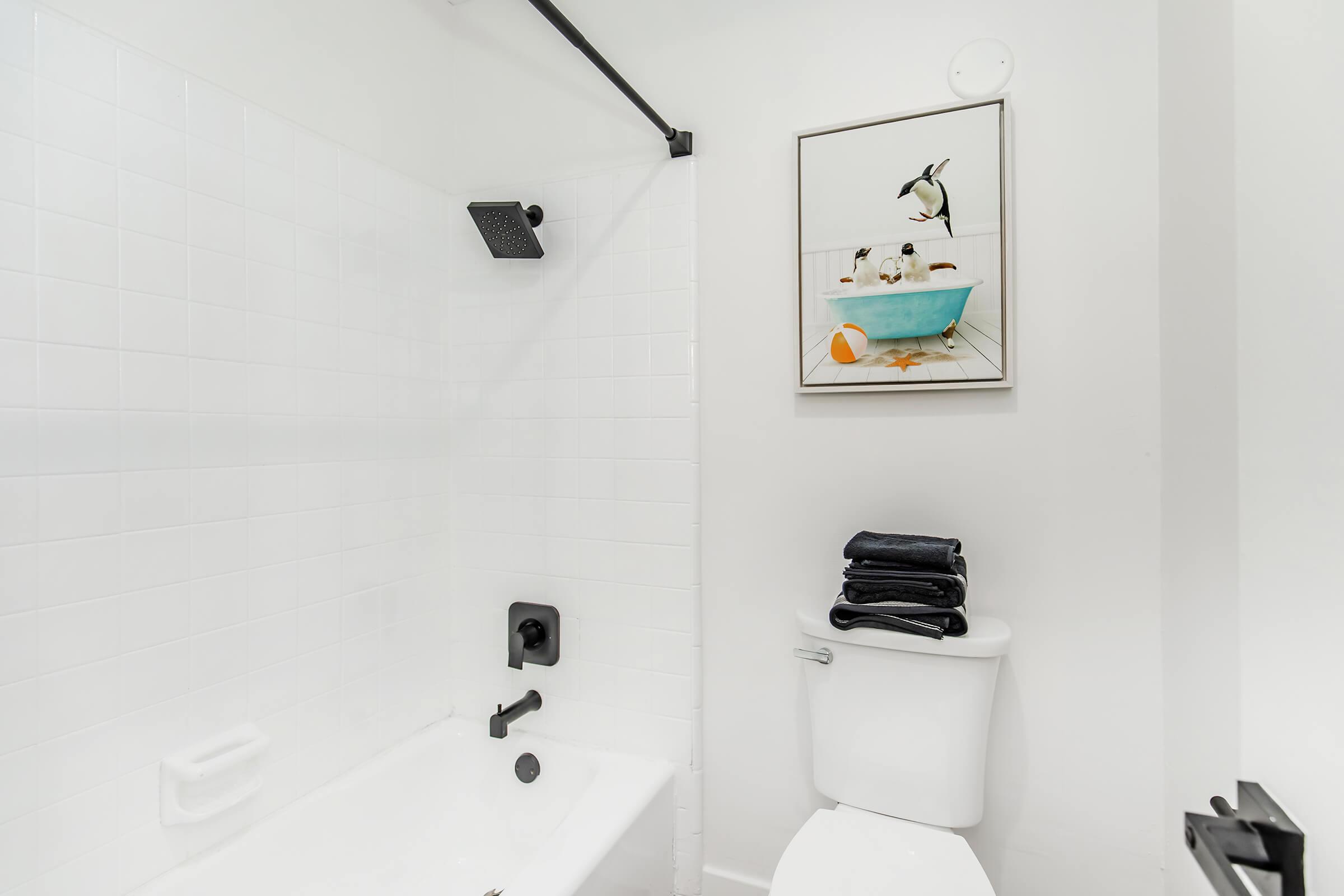 A bright, modern bathroom featuring a white tiled shower with a black showerhead, a white bathtub, a toilet, and a framed artwork of a penguin in a blue bathtub. Neatly stacked black towels are placed on the edge of the bathtub. The overall decor is clean and minimalist.