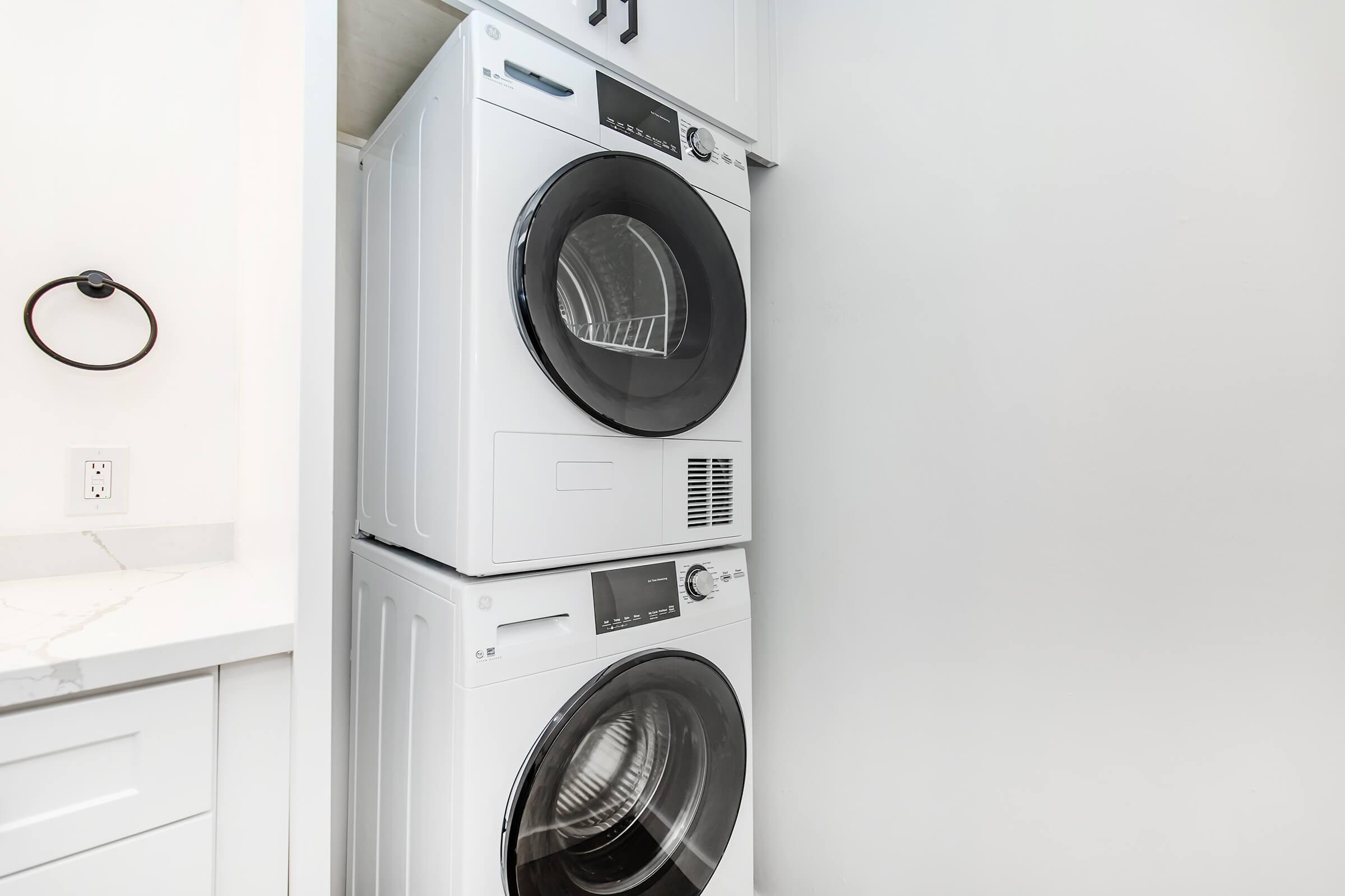 A stacked washer and dryer set in a compact laundry space. The appliances are white with black glass doors, positioned next to a light-colored countertop and a wall. A towel holder is mounted on the wall nearby, with a minimalistic, clean design. The overall setting is bright and modern.